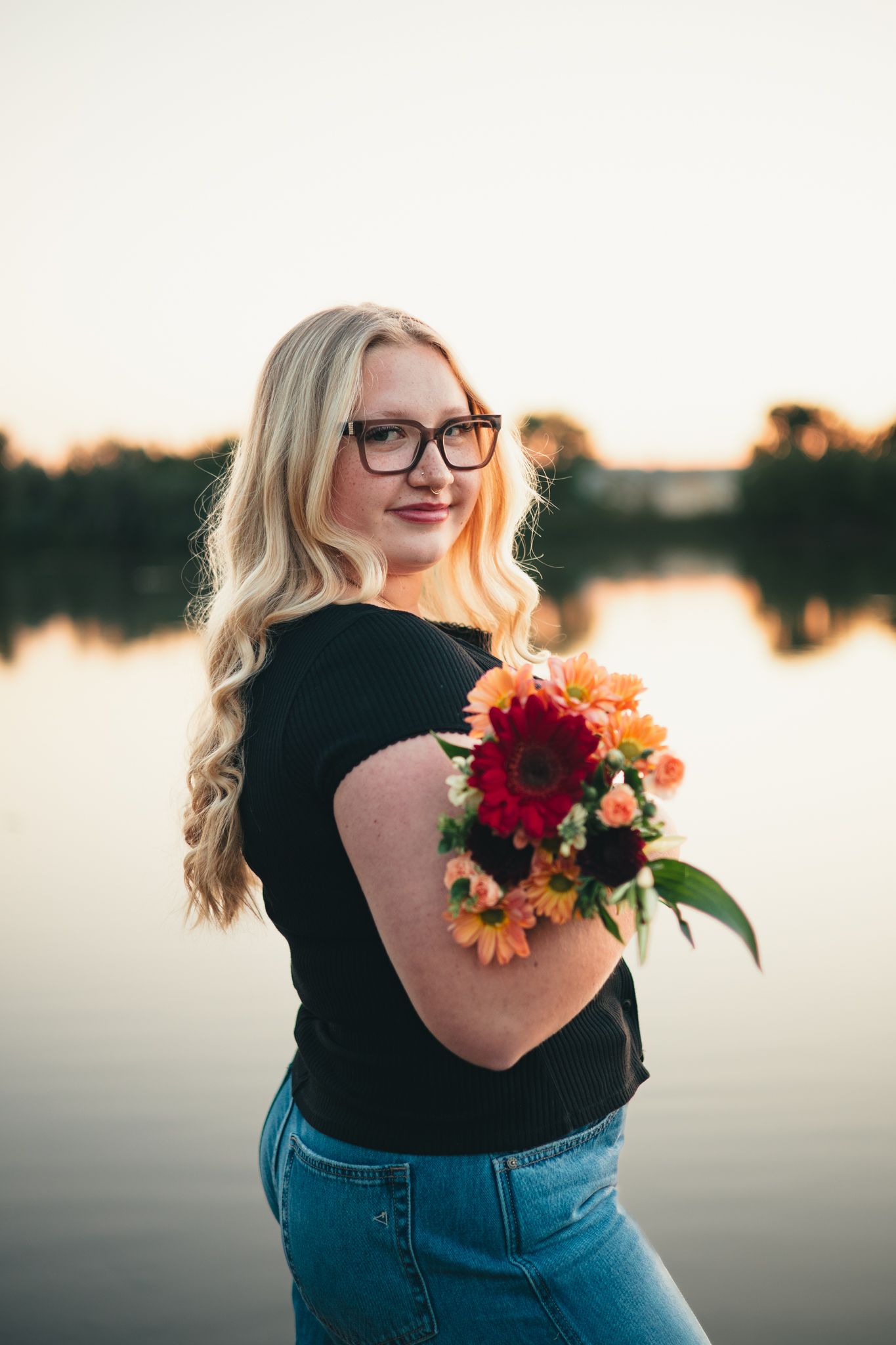 A senior in a black top and jeans holds vibrant red and orange flowers against a sunset-lit lake backdrop. photo, Great Falls, MT
