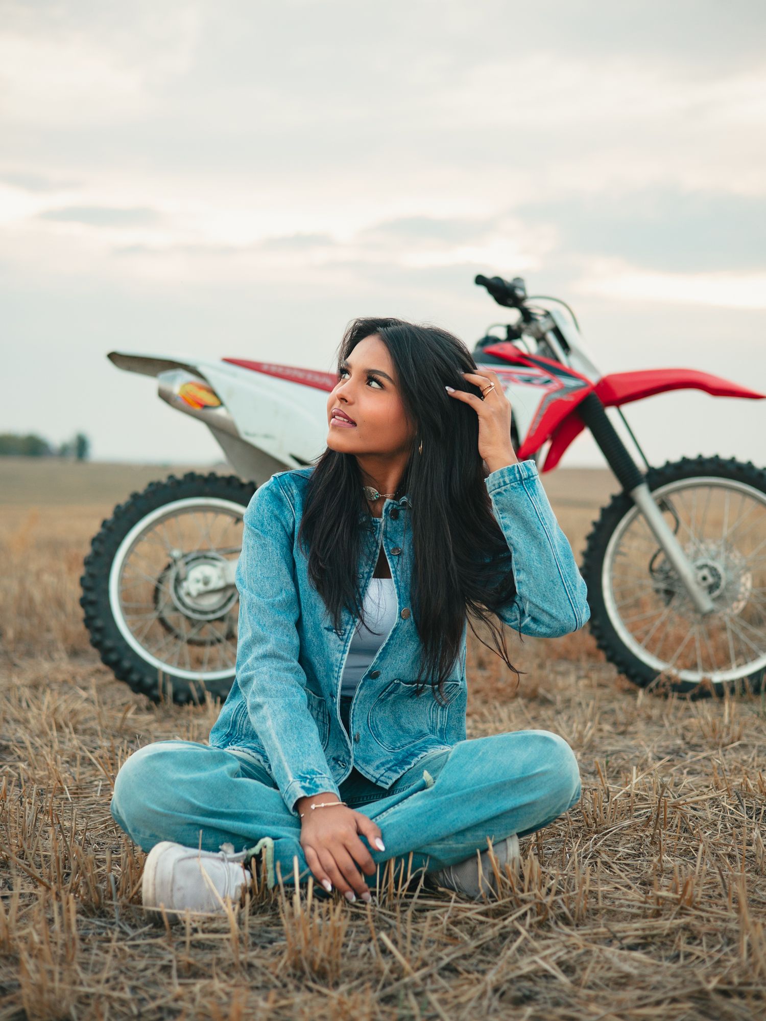 A senior in denim sits casually on dry grass next to a red dirt bike on an overcast day. photo, Great Falls, MT