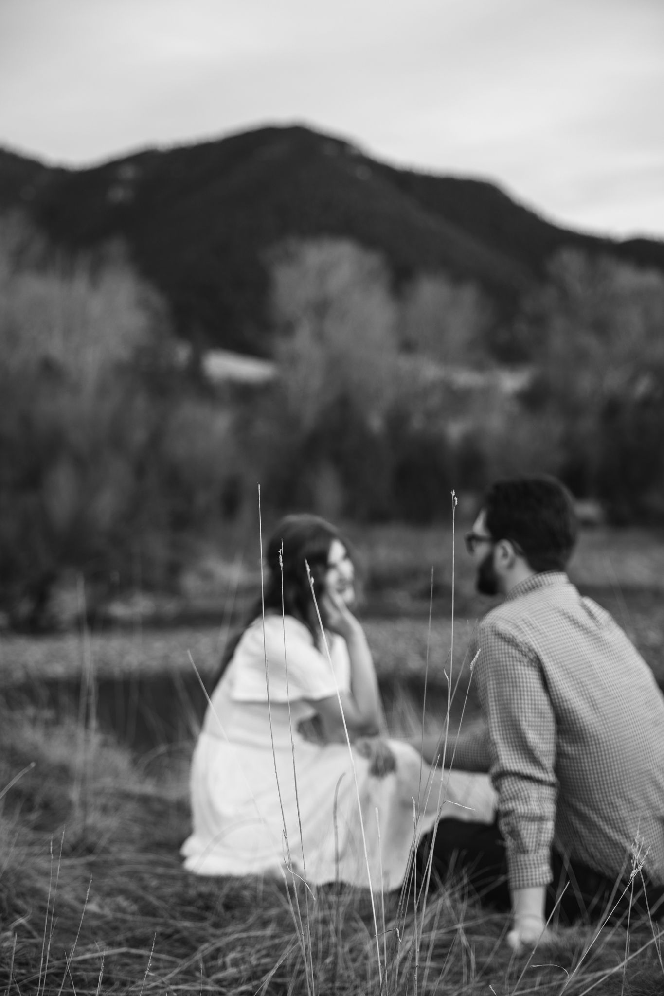 Black and white photo of a couple sitting in a field with mountains and trees in the background.