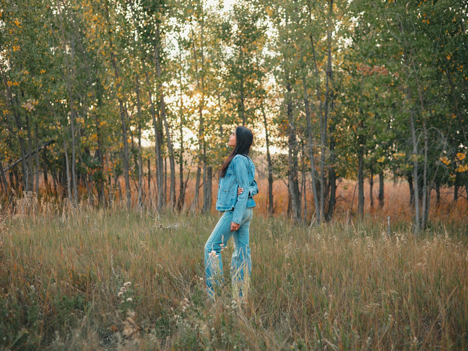 senior in light blue dress walks through tall grass in a golden-lit forest clearing at sunset. photo, Great Falls, MT