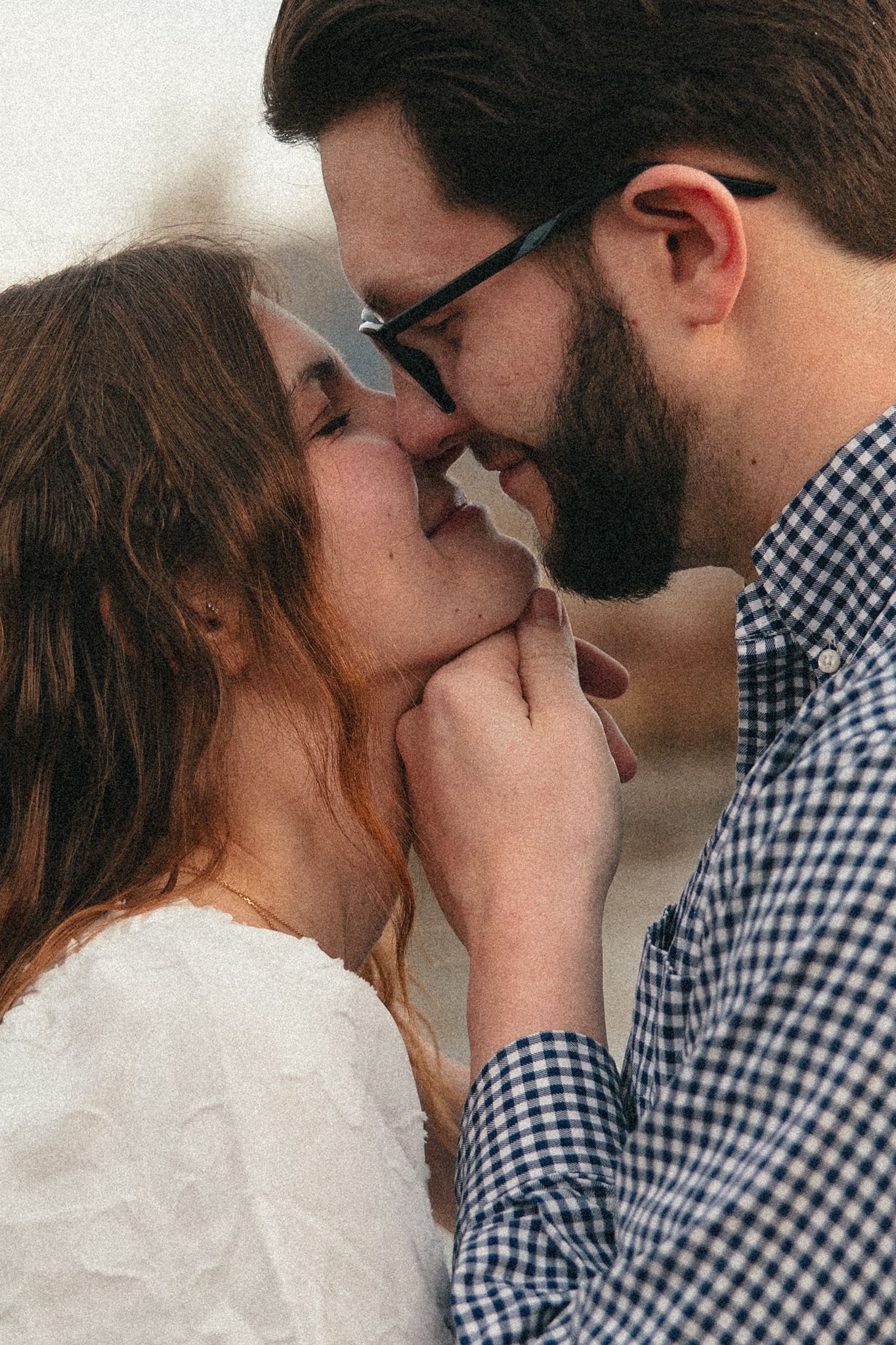 A romantic couple sharing an intimate moment, the bearded man in glasses and plaid shirt tenderly holds the woman's face.