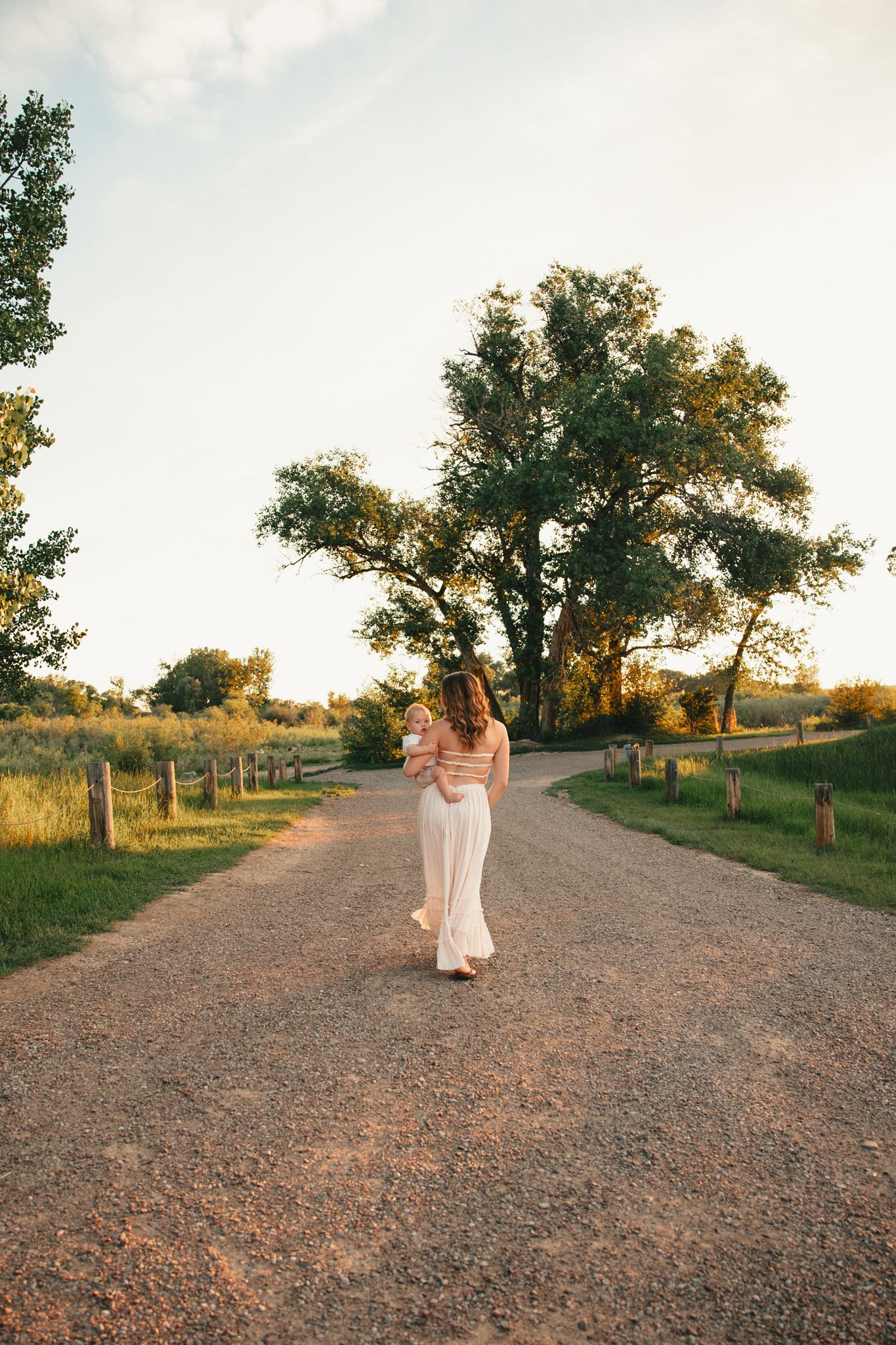 Woman in white flared pants and striped top walking down a scenic gravel path surrounded by lush green trees at sunset.