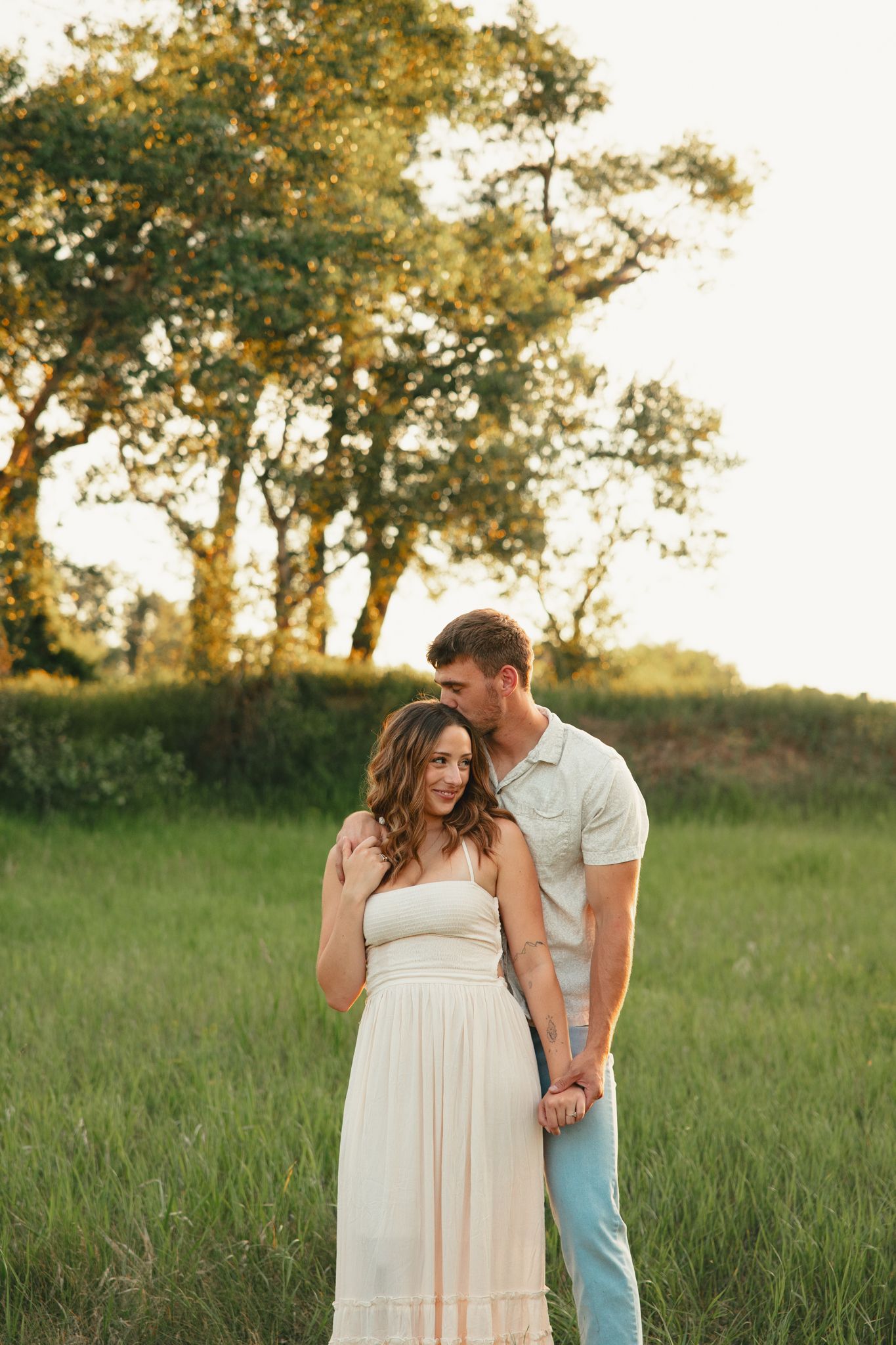 A couple embraces in a sunlit green field, woman in white maxi dress, man in light blue outfit, golden trees behind them.