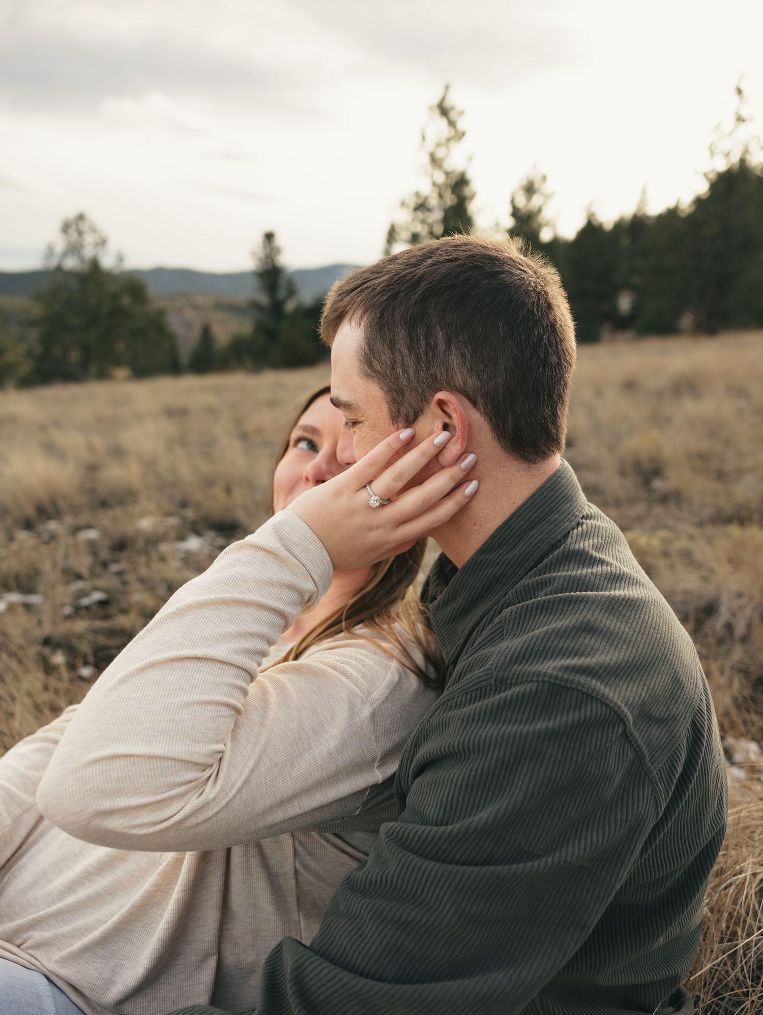 Couple sitting in a field, woman wearing engagement ring touching man's face, with trees and mountains in background.