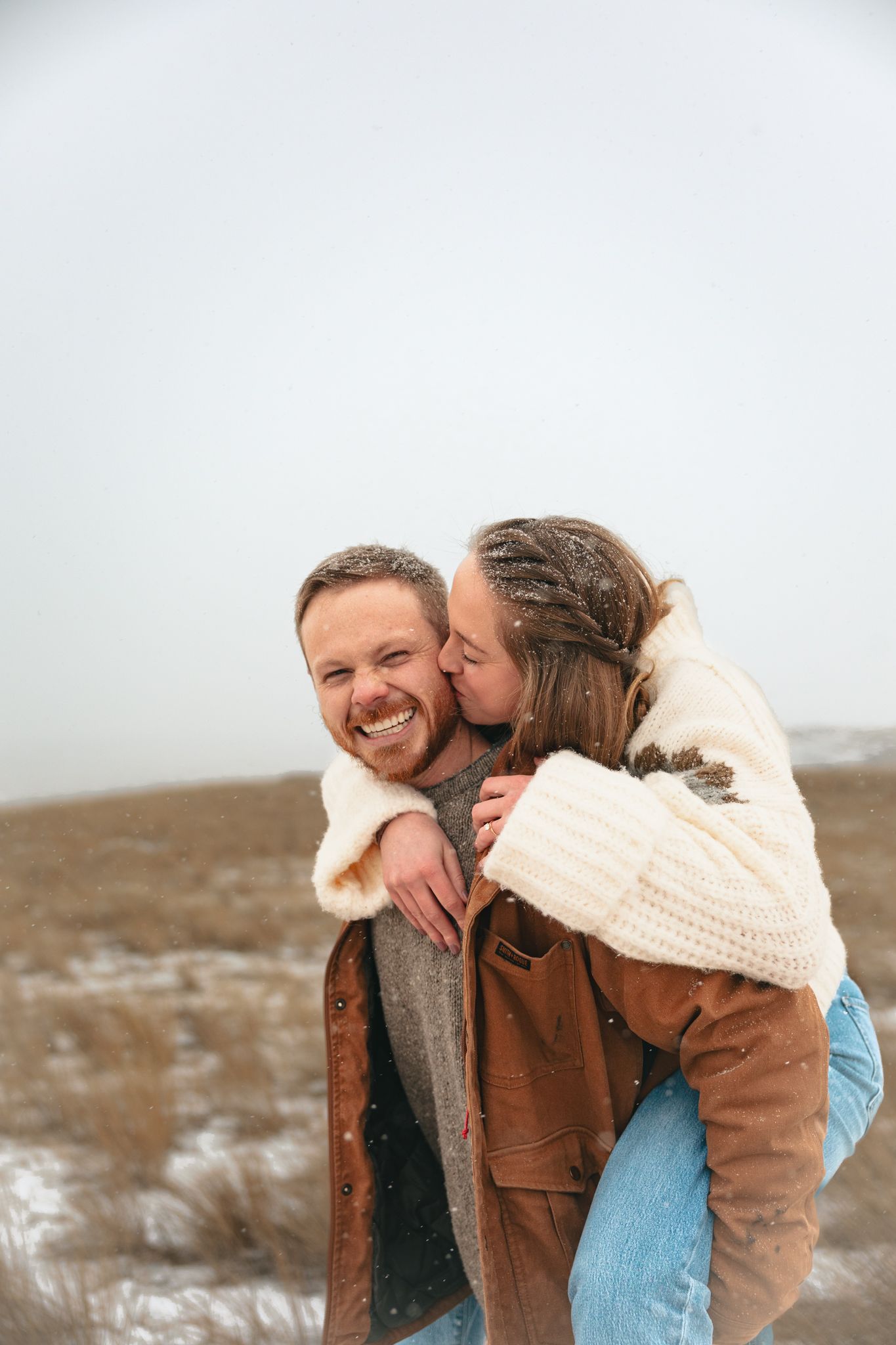 A happy couple shares a piggyback ride in a snowy field, with the woman kissing the smiling man on the cheek.