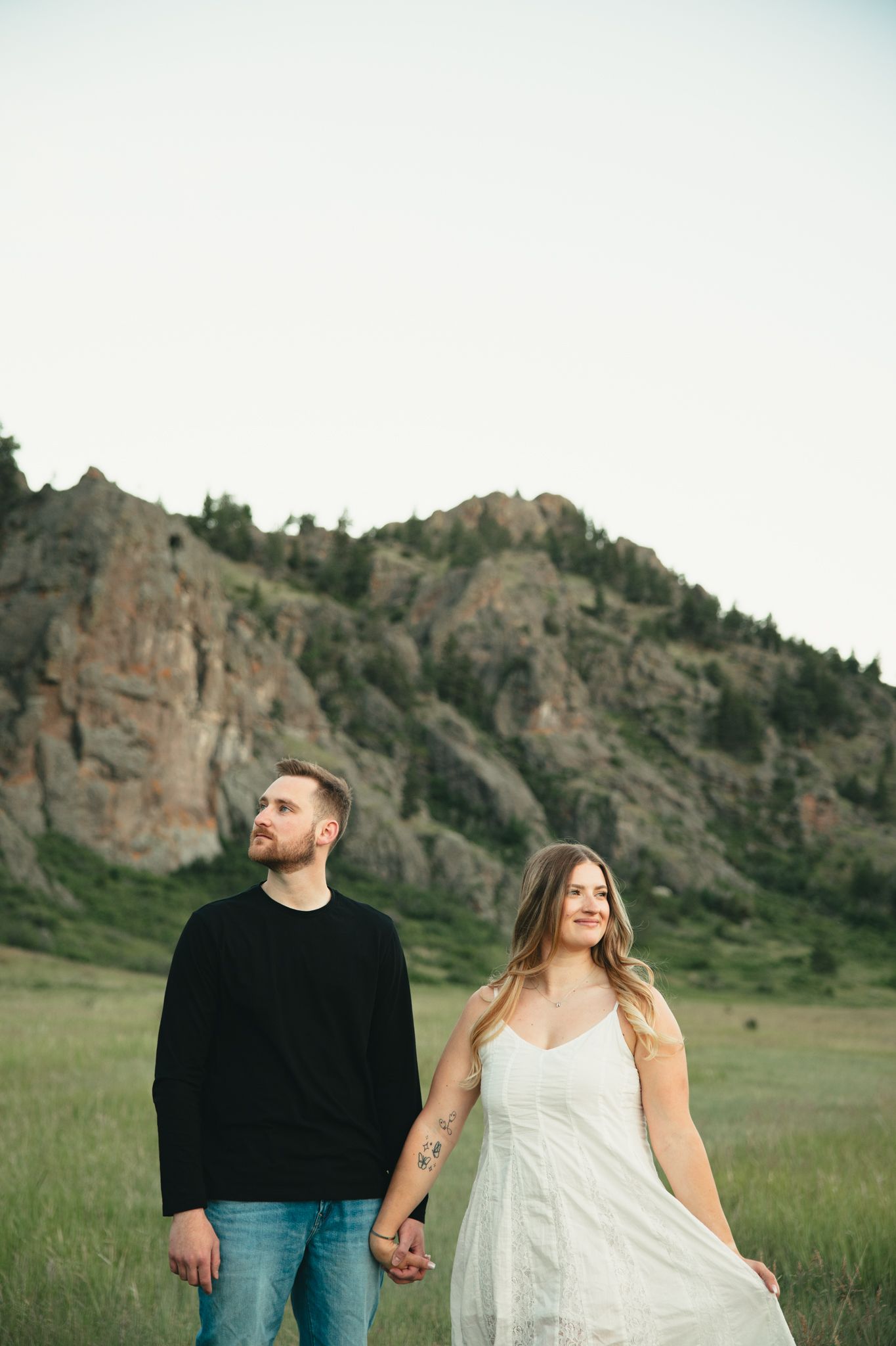Couple holding hands in a green meadow with rocky mountain backdrop, woman in white dress, man in black sweater.