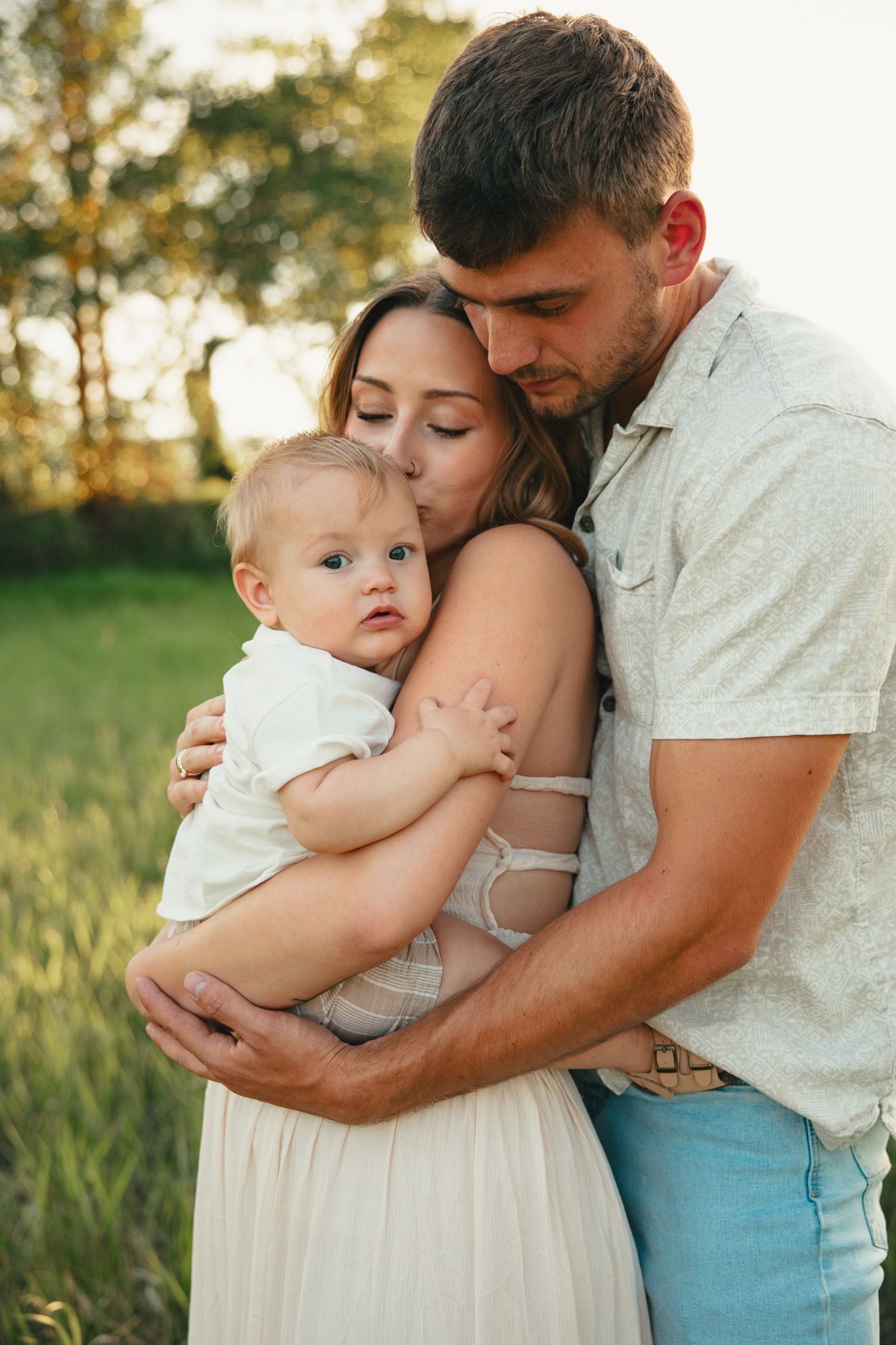 Young family outdoors at sunset, parents embracing baby in white outfit in a green field.