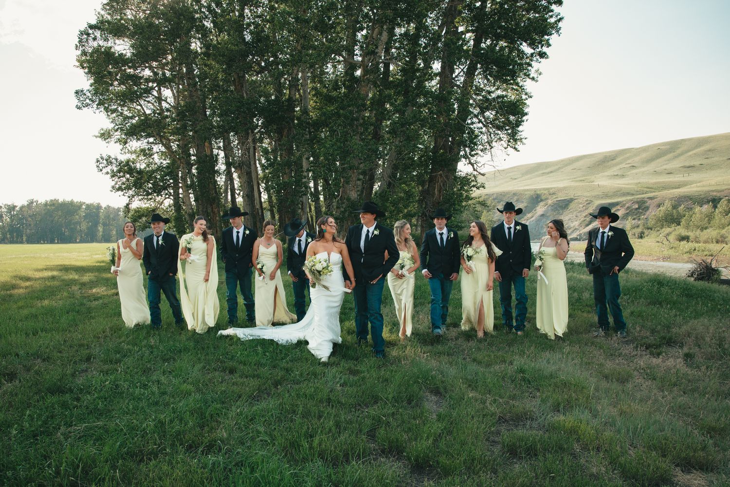 Wedding party in yellow dresses and suits posing on green grass with trees and rolling hills in background.