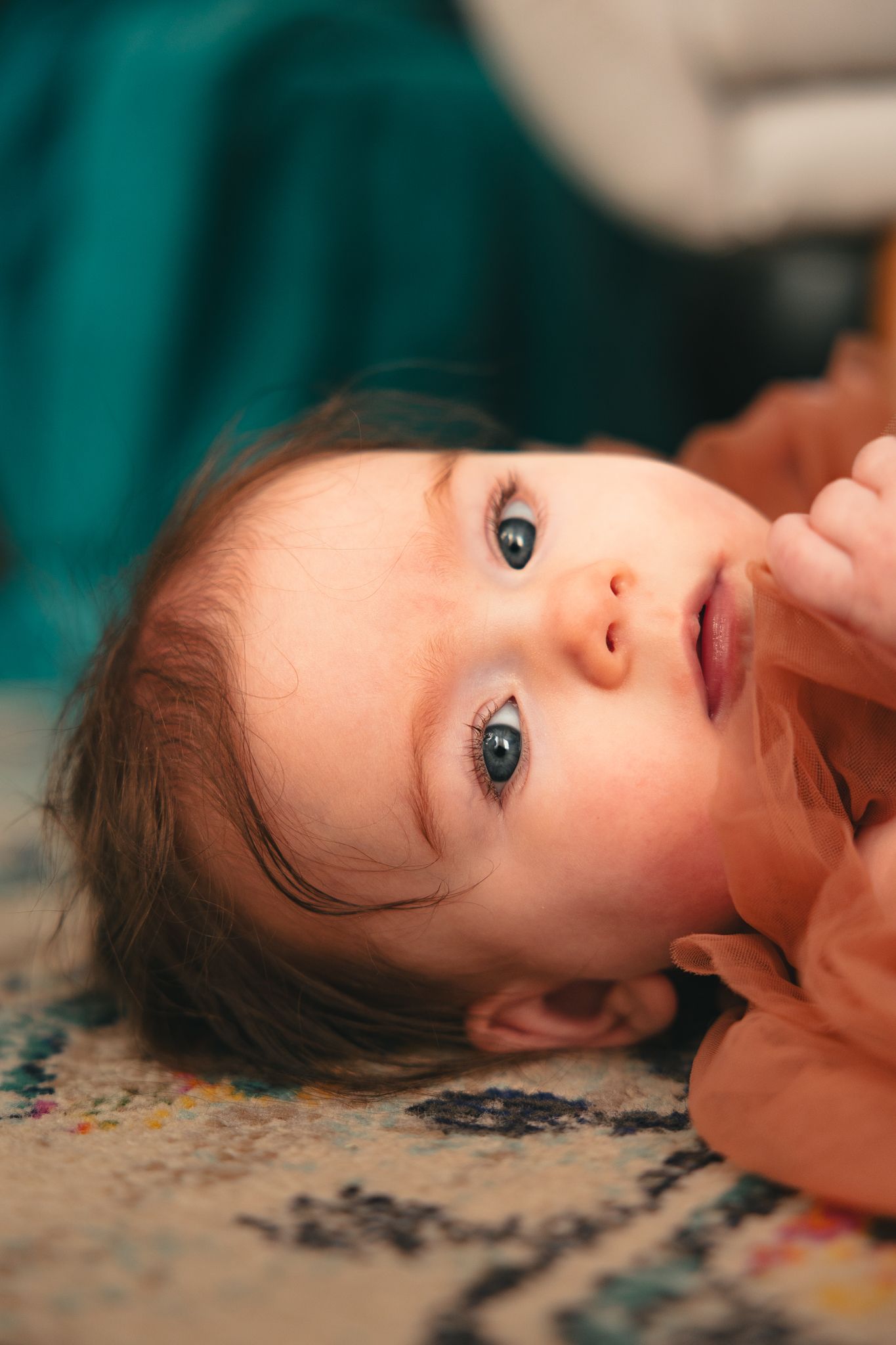 Adorable baby with blue eyes lying on a patterned rug, wearing an orange outfit, looking up curiously.