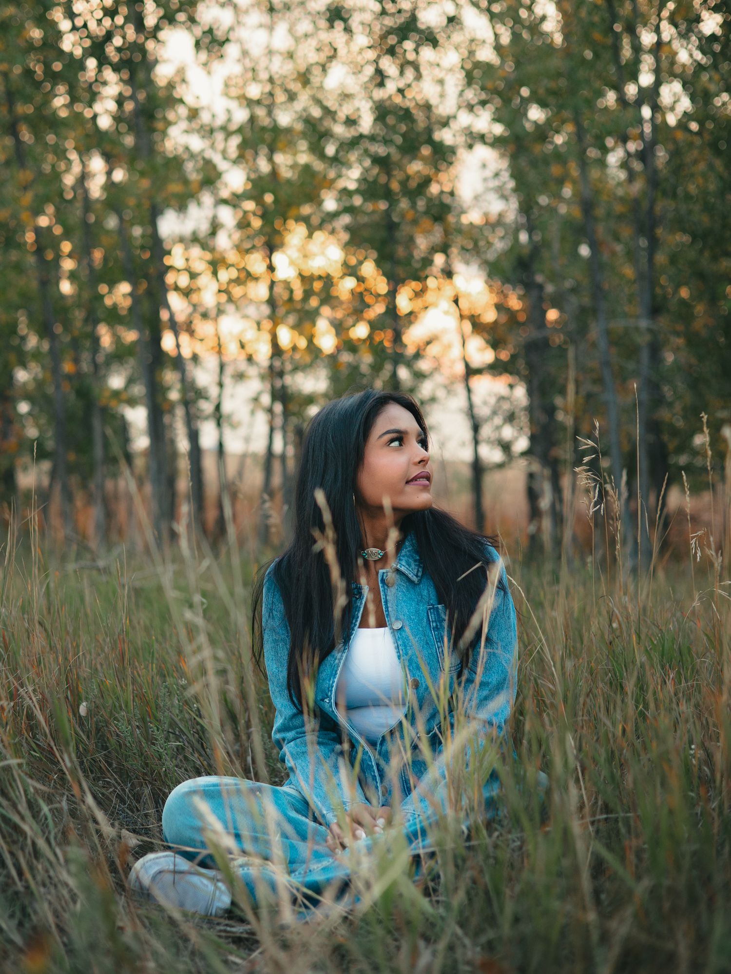 A senior in a blue denim jacket poses thoughtfully in a field of tall grass during golden hour sunset. photo, Great Falls, MT
