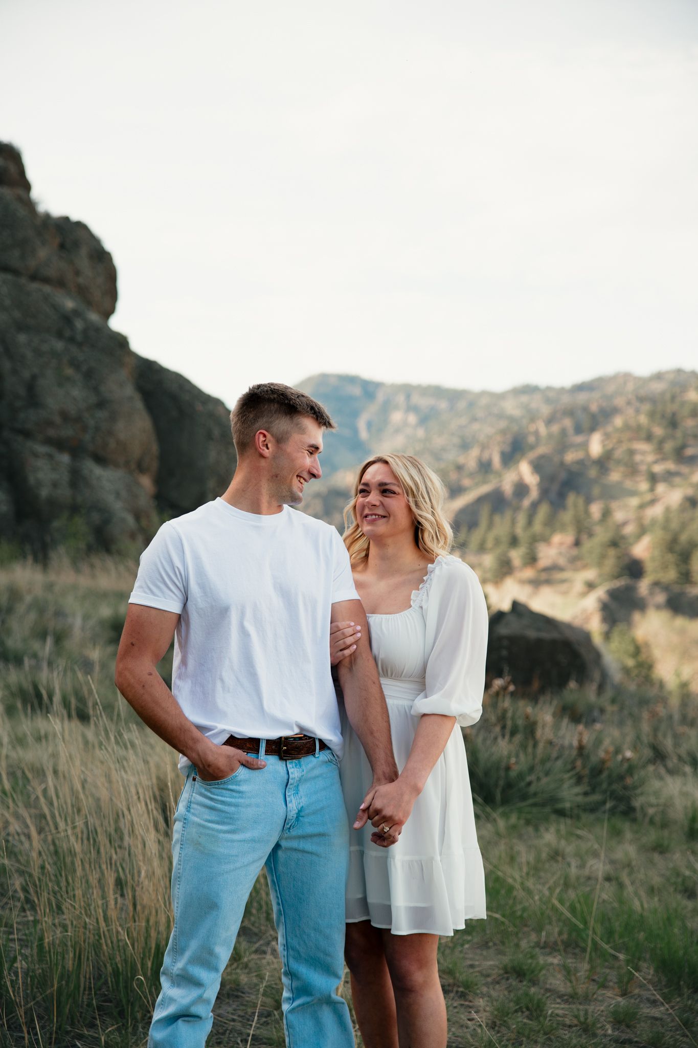 A happy couple in white outfits holds hands while smiling at each other in a scenic mountain landscape.