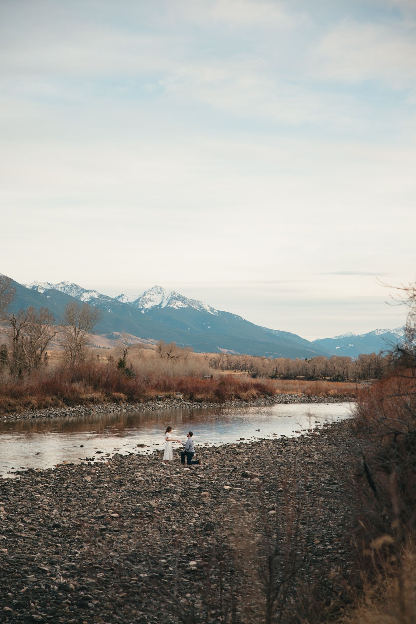 Couple standing on rocky riverbank with snow-capped mountains and autumn trees in background.