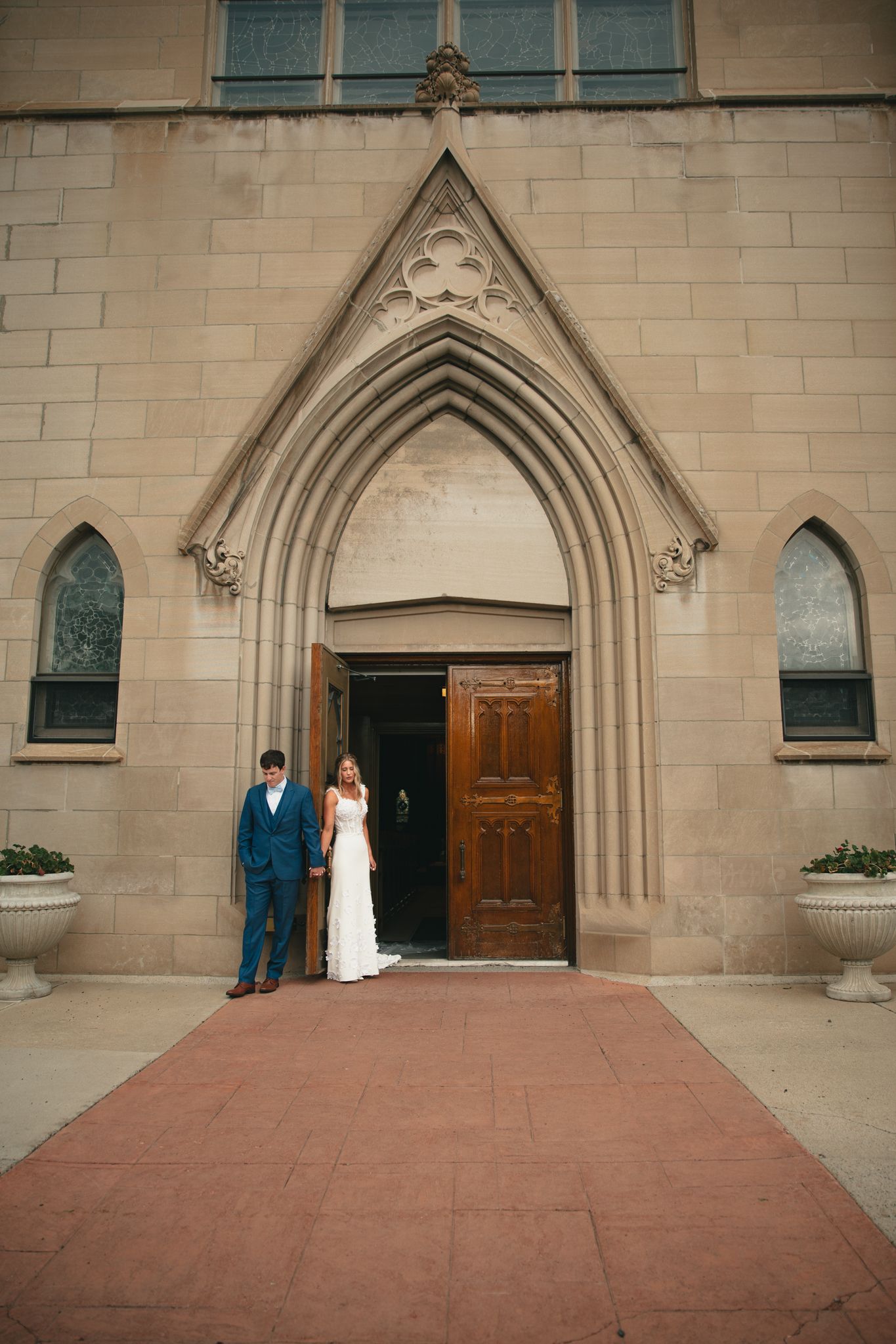Bride and groom standing at ornate Gothic church entrance with wooden doors and red brick pathway.