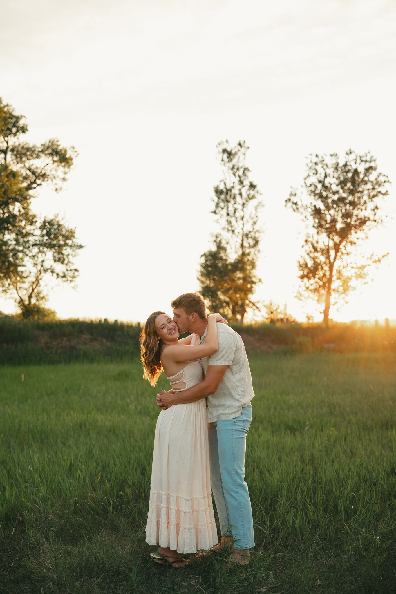 Couple embracing in a sunlit green field at golden hour, woman in white dress, romantic outdoor portrait.
