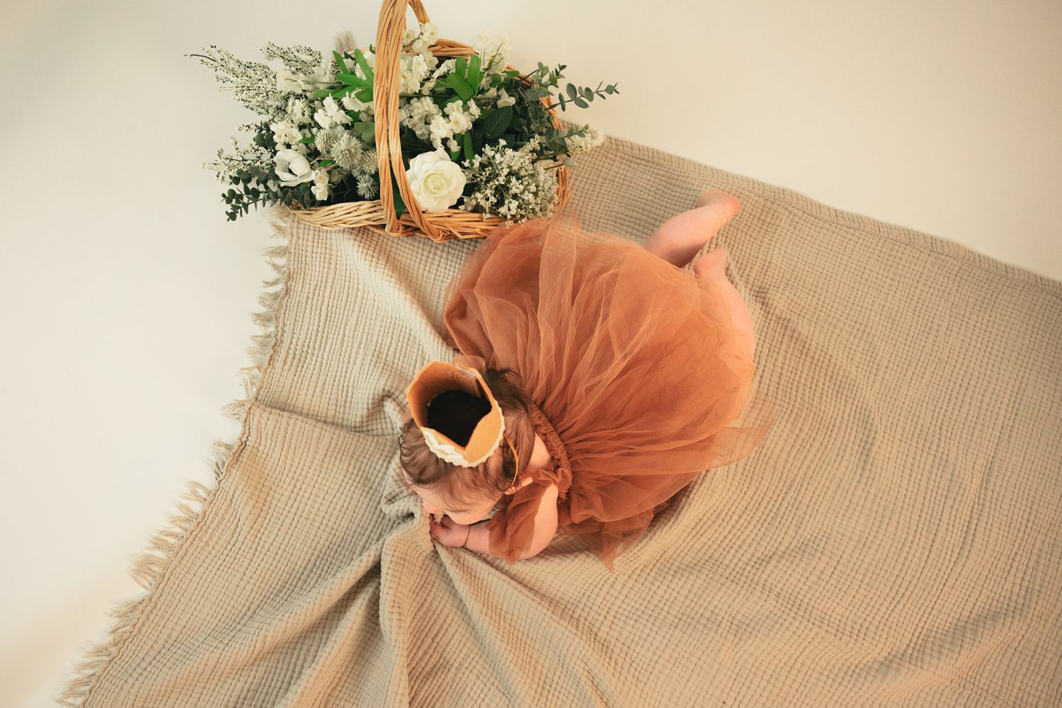 Baby in rust tulle dress lying on beige linen blanket beside a wicker basket of white flowers.