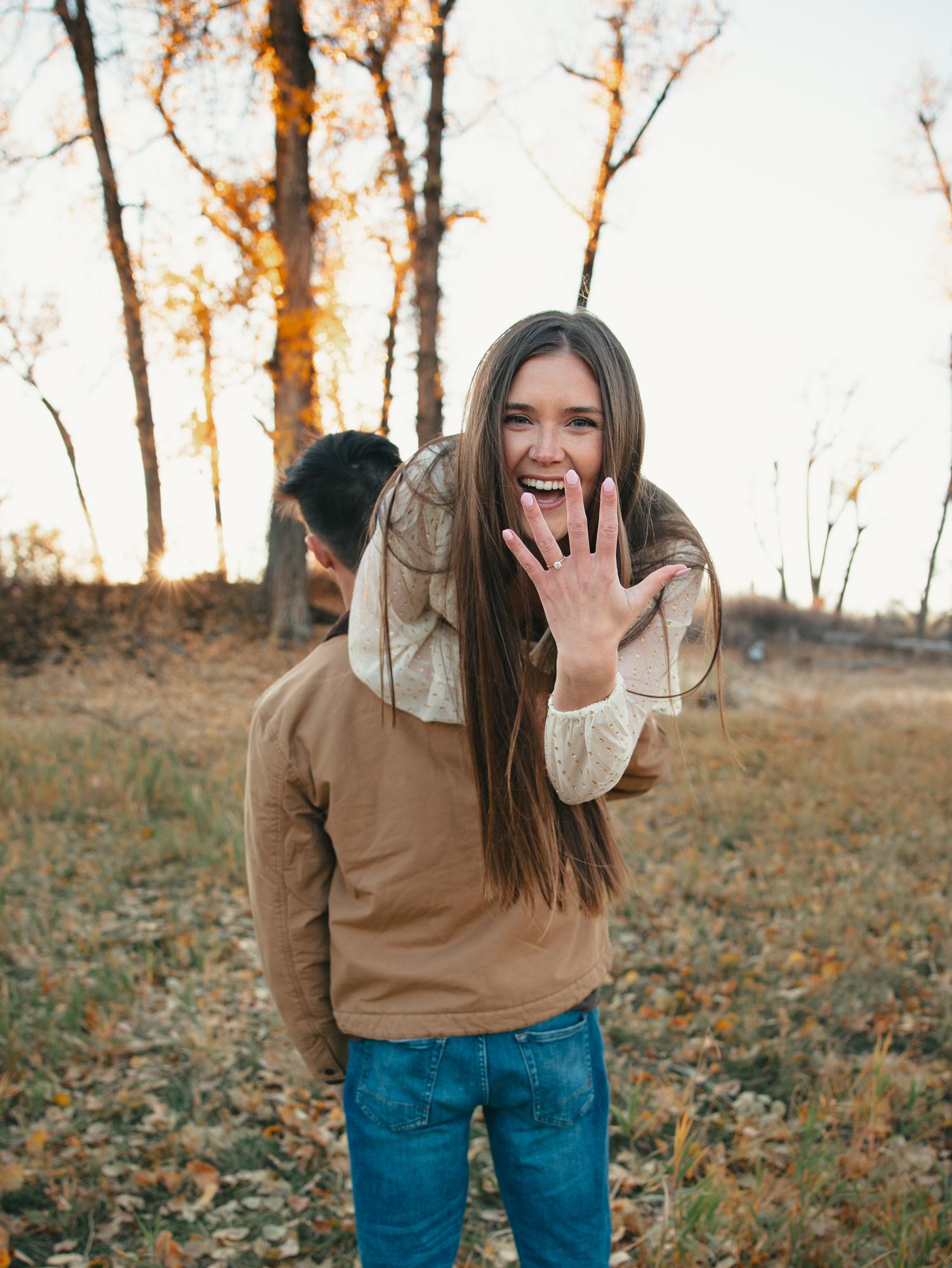 Woman showing off engagement ring while embracing partner outdoors in autumn setting with fall foliage.