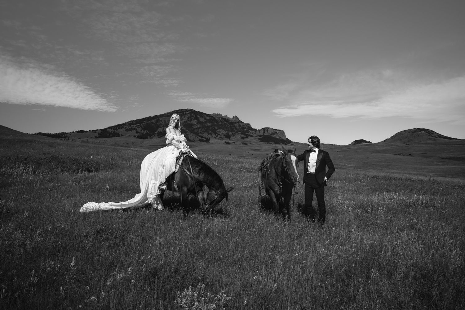 Black and white photo of a bride and groom with horses in a vast grassy field with hills in the background.