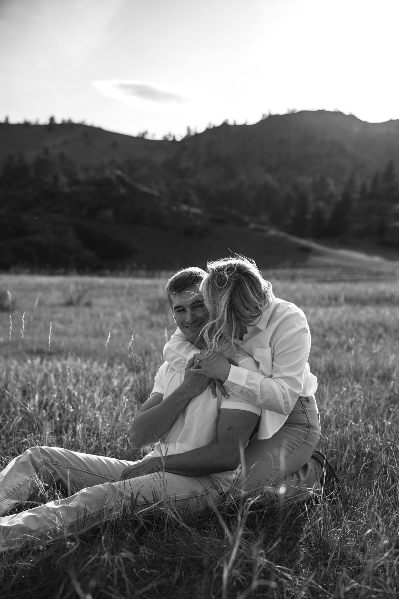 Black and white photo of a couple sitting in a grassy field, embracing warmly with mountains and trees in the background.