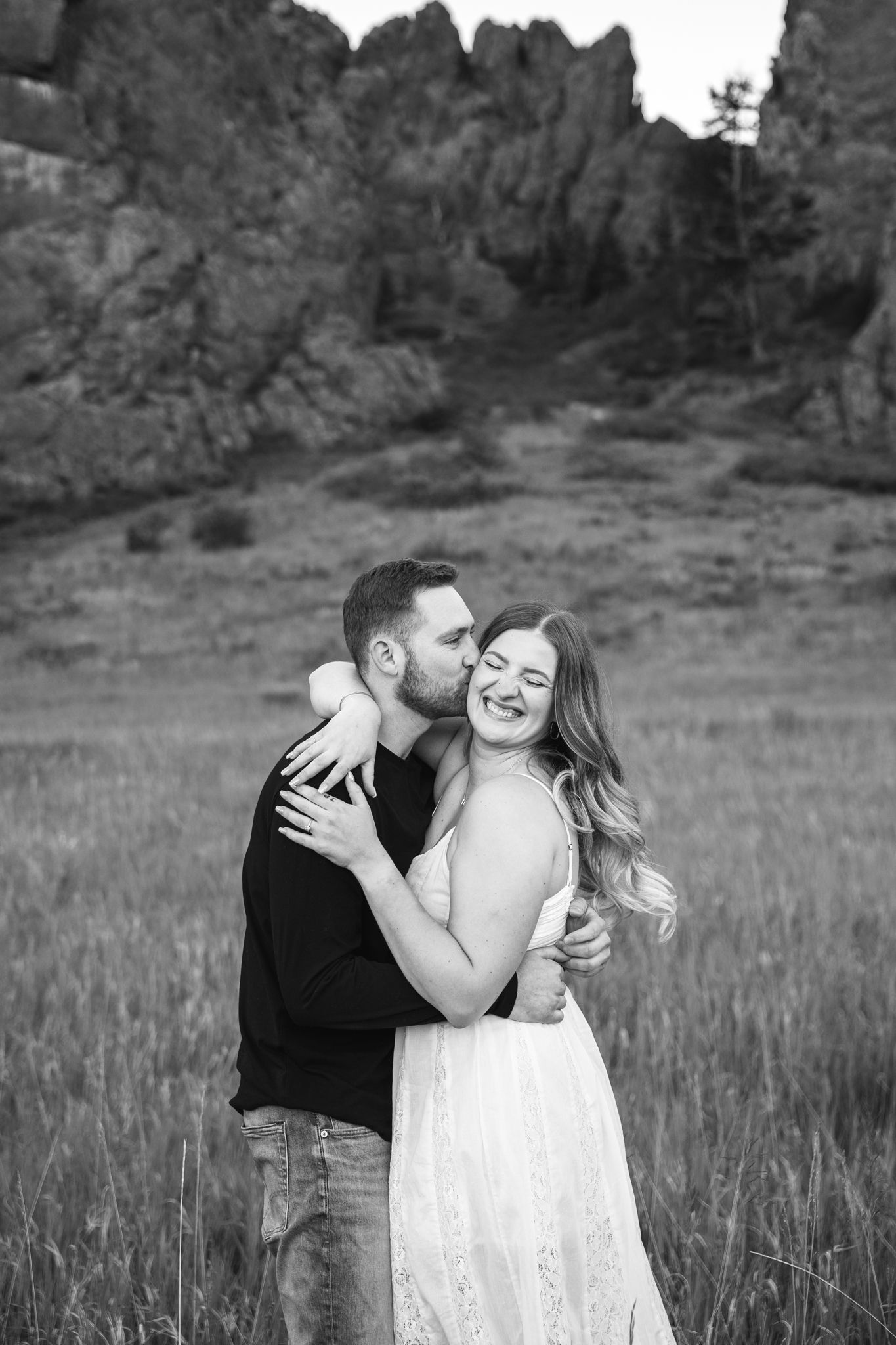 Black and white photo of a couple embracing in a field, man kissing woman's cheek, rocky mountains in background.
