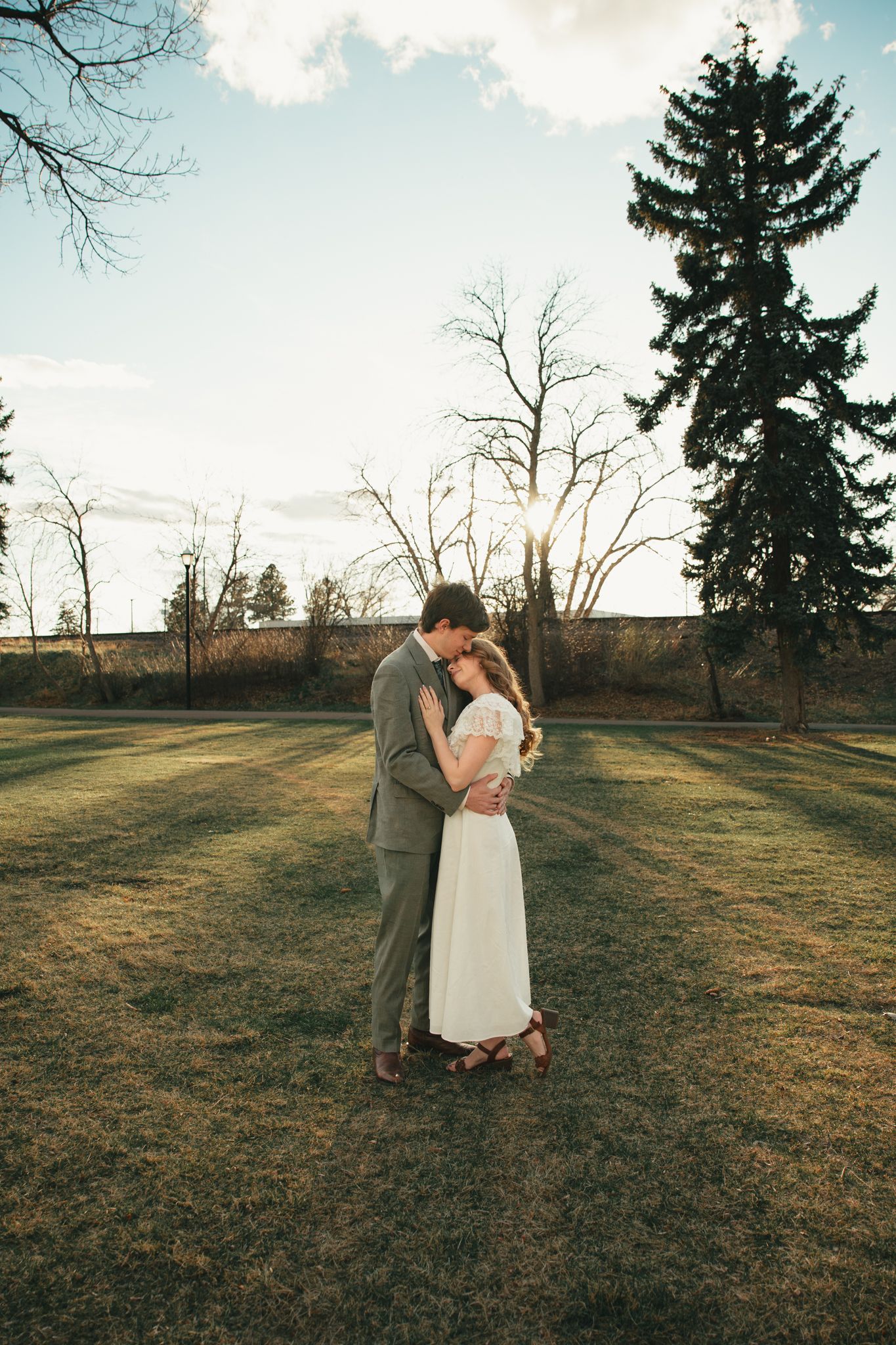Couple embracing in golden hour light on green lawn, groom in gray suit, bride in white dress, evergreen tree backdrop.