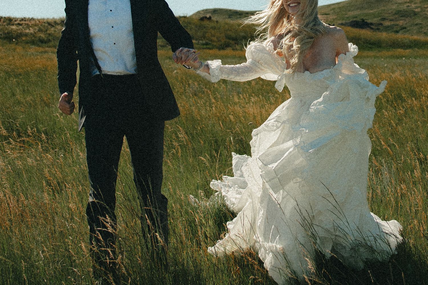 A bride in a flowing white gown and groom in black suit hold hands running through a lush green field.