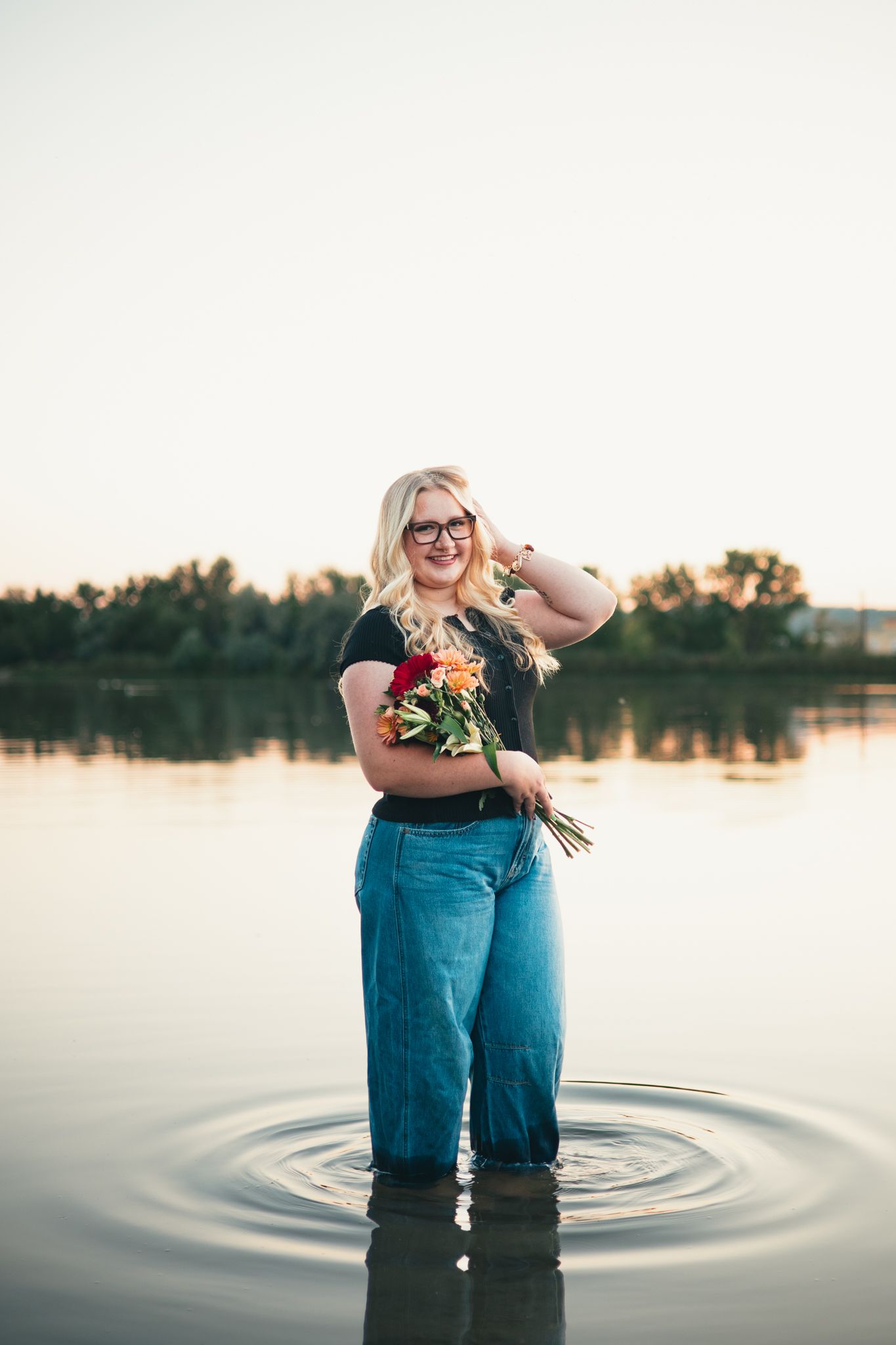 senior wearing glasses and jeans stands in shallow lake water at sunset holding flowers while creating ripples. photo, Great Falls, MT
