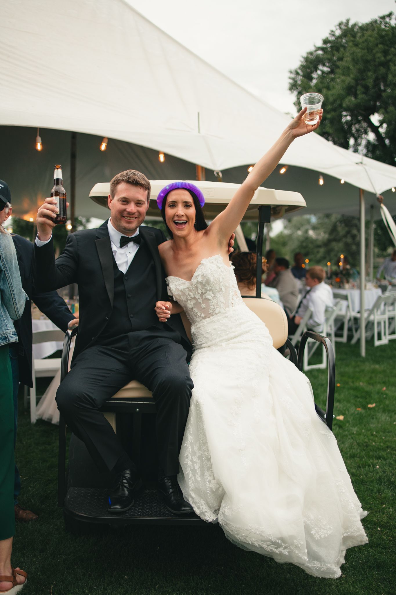 Joyful bride in white lace gown and purple hair raises glass beside groom in tuxedo on golf cart at outdoor wedding reception.