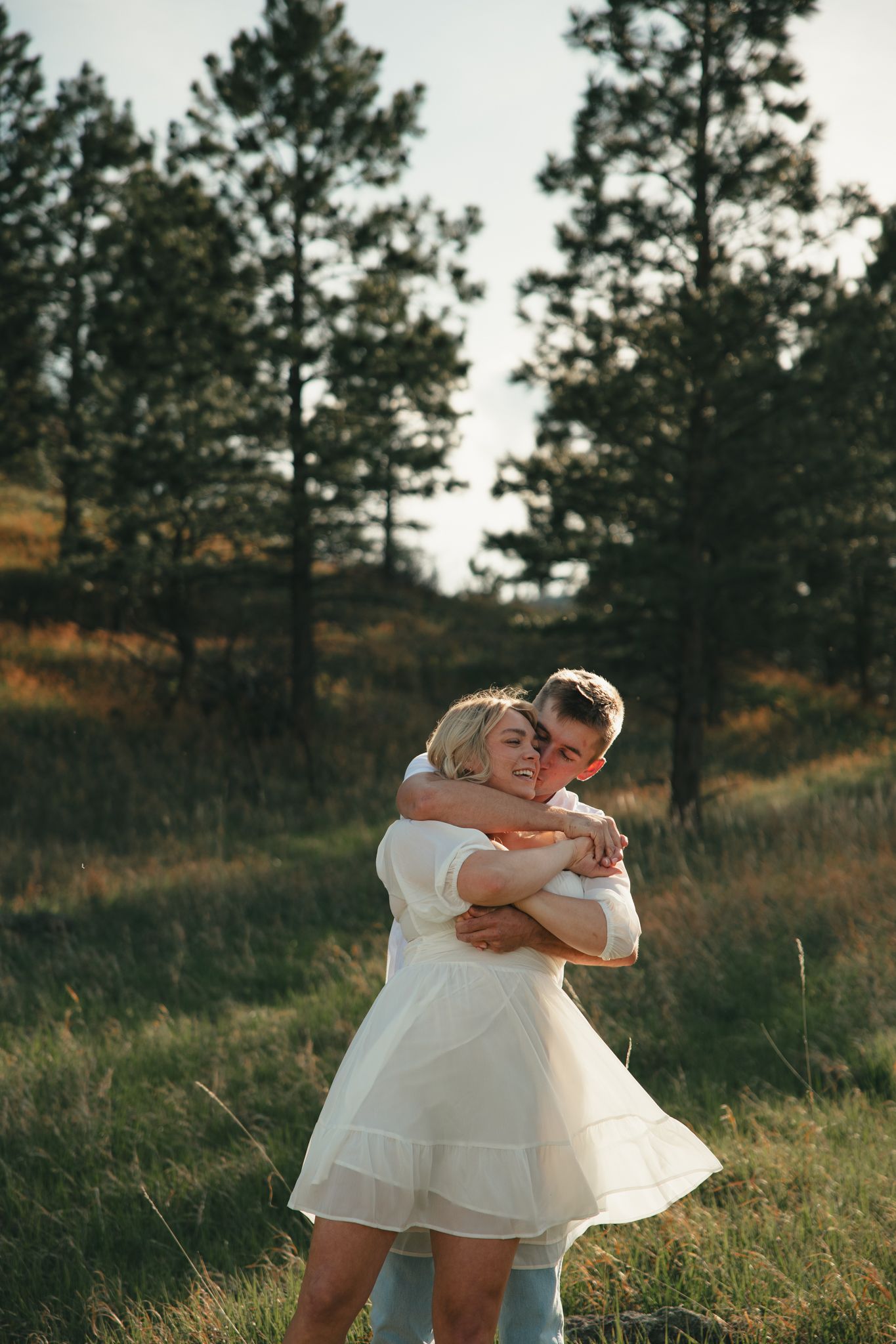 Couple embracing in a sunlit meadow with pine trees, woman in white dress, romantic outdoor photography.