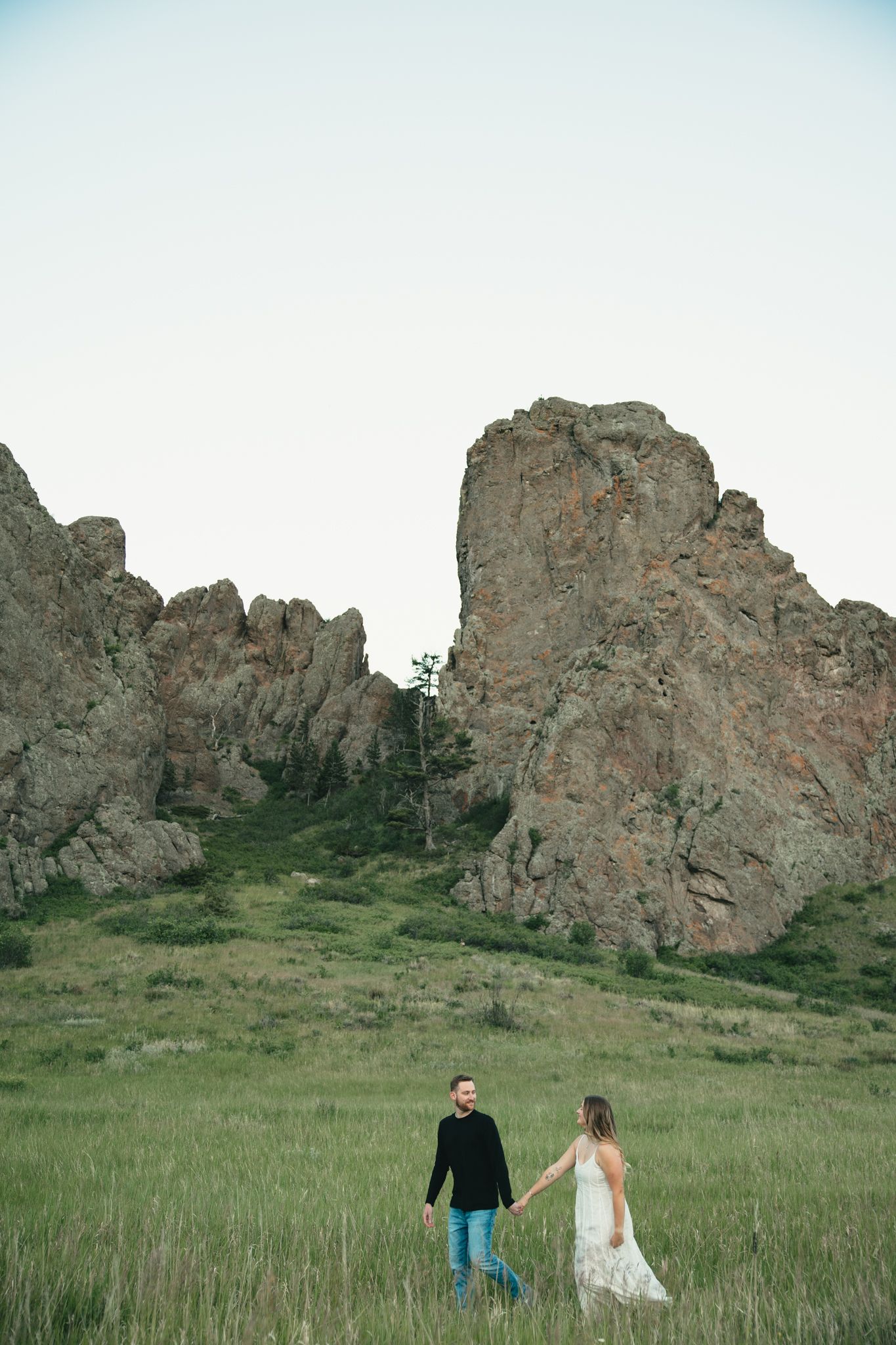 Couple holding hands in a green meadow with dramatic rocky cliffs in the background during an outdoor engagement session.