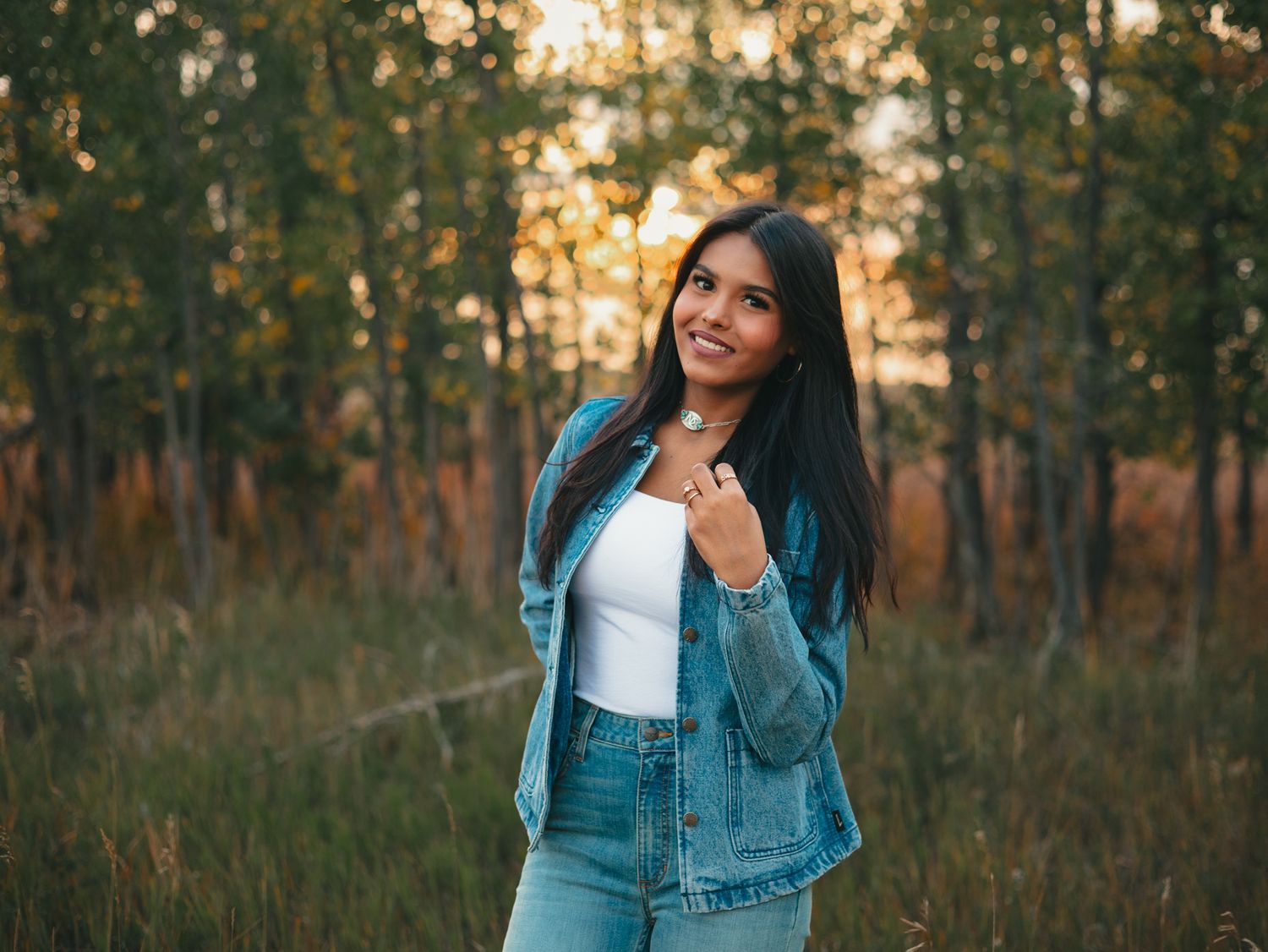 A woman in a denim jacket poses outdoors during golden hour in a field with trees in the background. senior photo, Great Falls, Montana