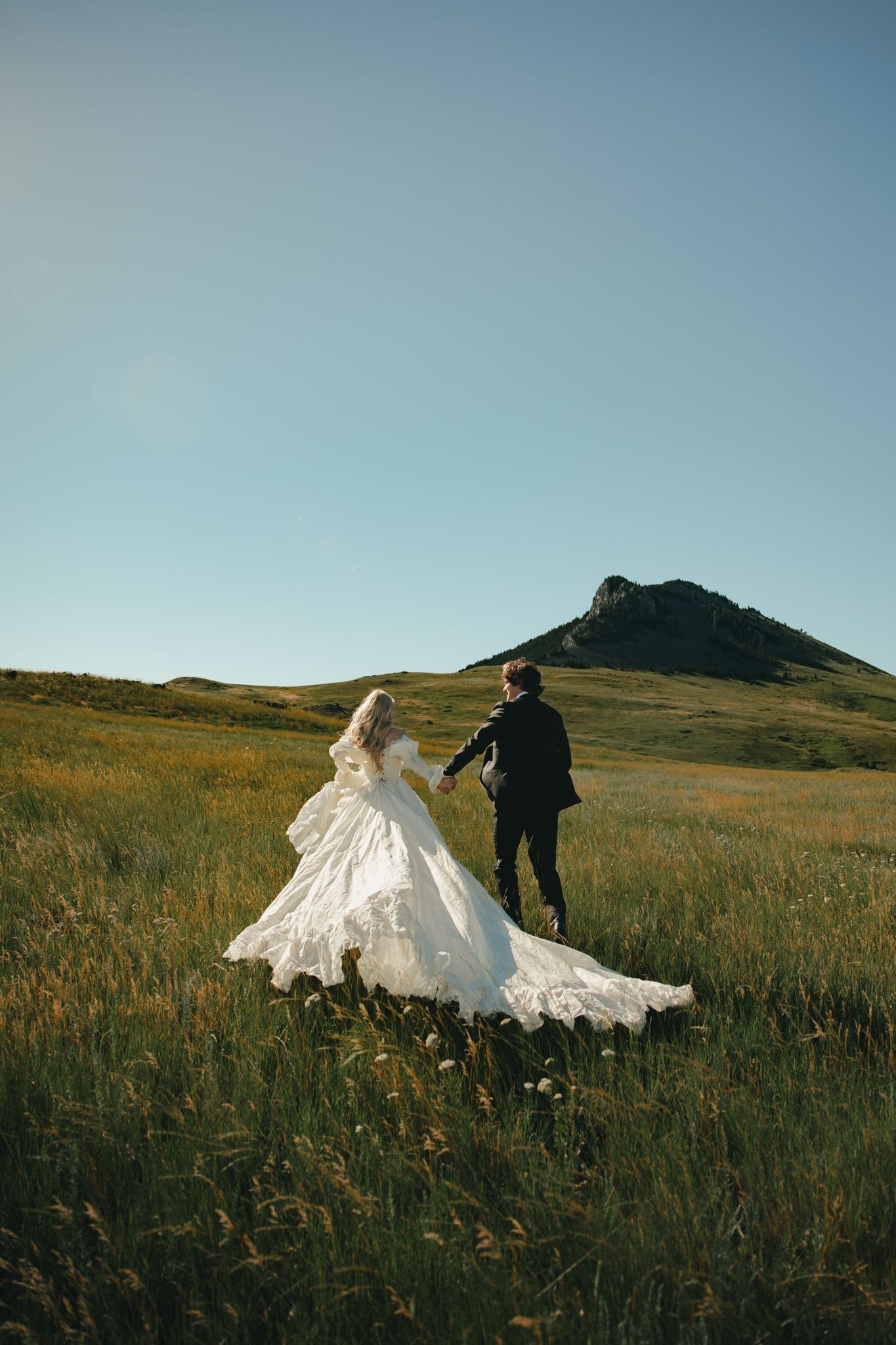 Bride and groom running through green meadow with mountain backdrop on sunny wedding day.