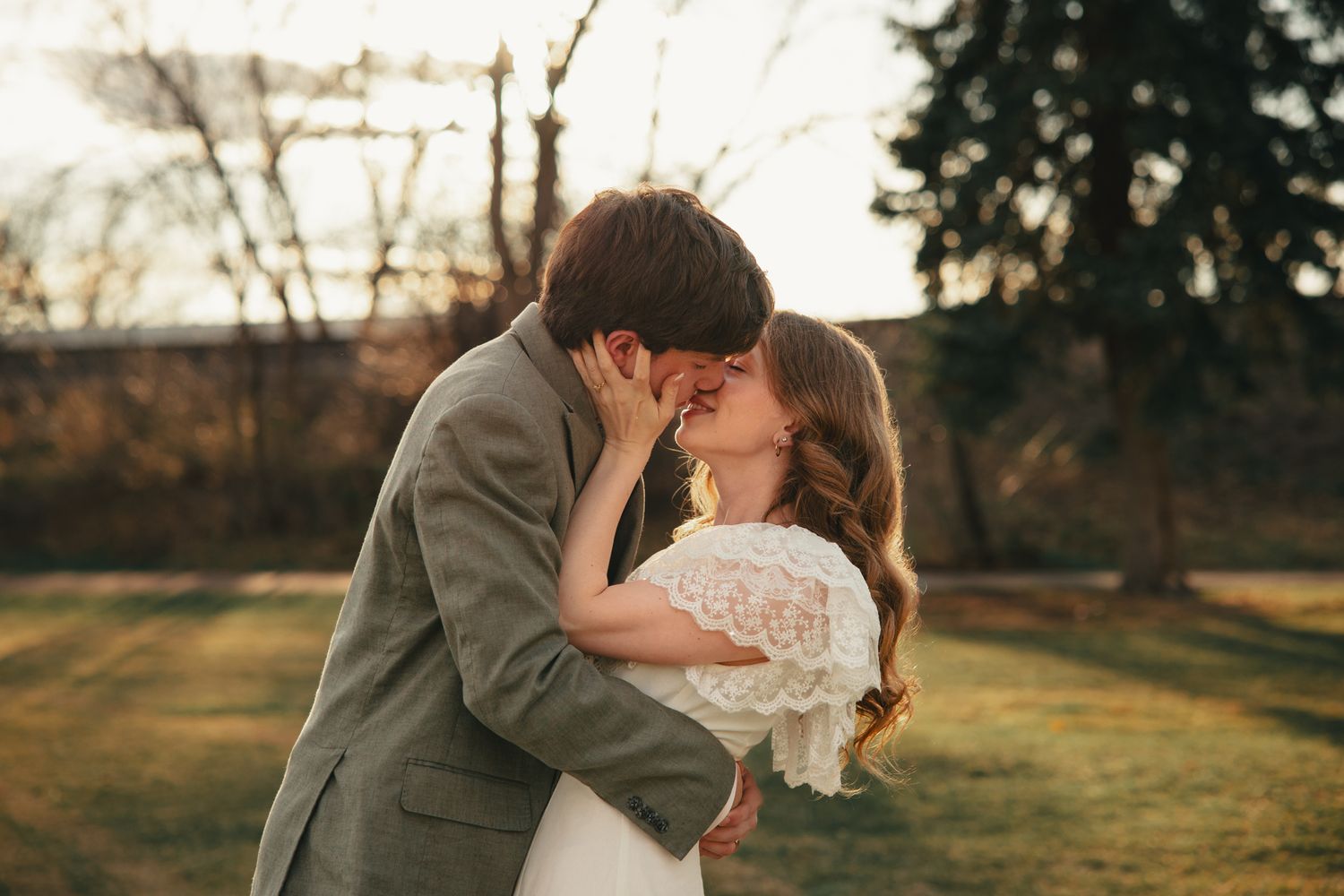 A couple shares a romantic kiss outdoors during golden hour, bride holding bouquet in white lace dress, groom in gray suit.