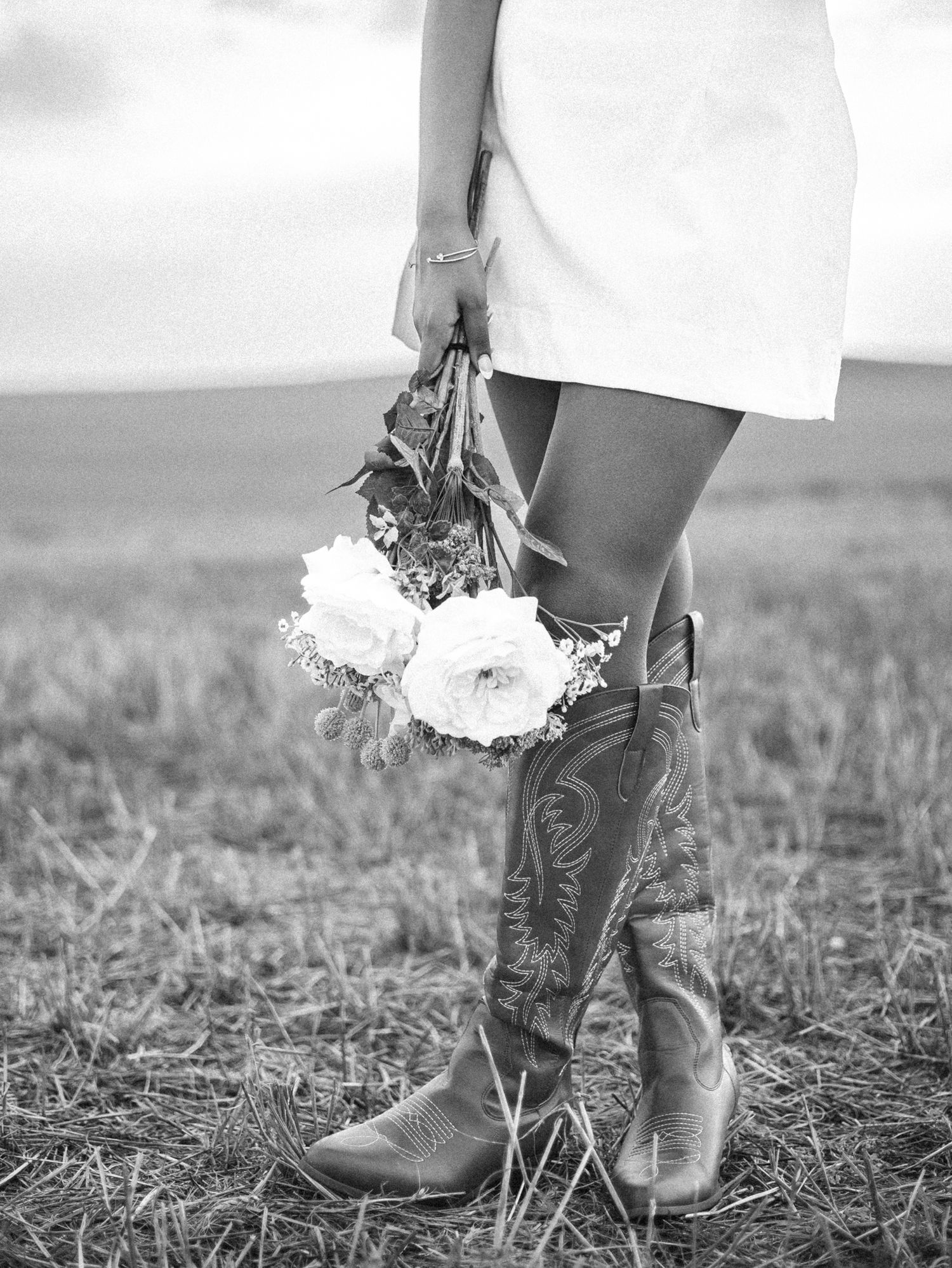 Black and white image shows a person wearing boots holding white flowers while standing in a grassy field. photo, Great Falls, MT