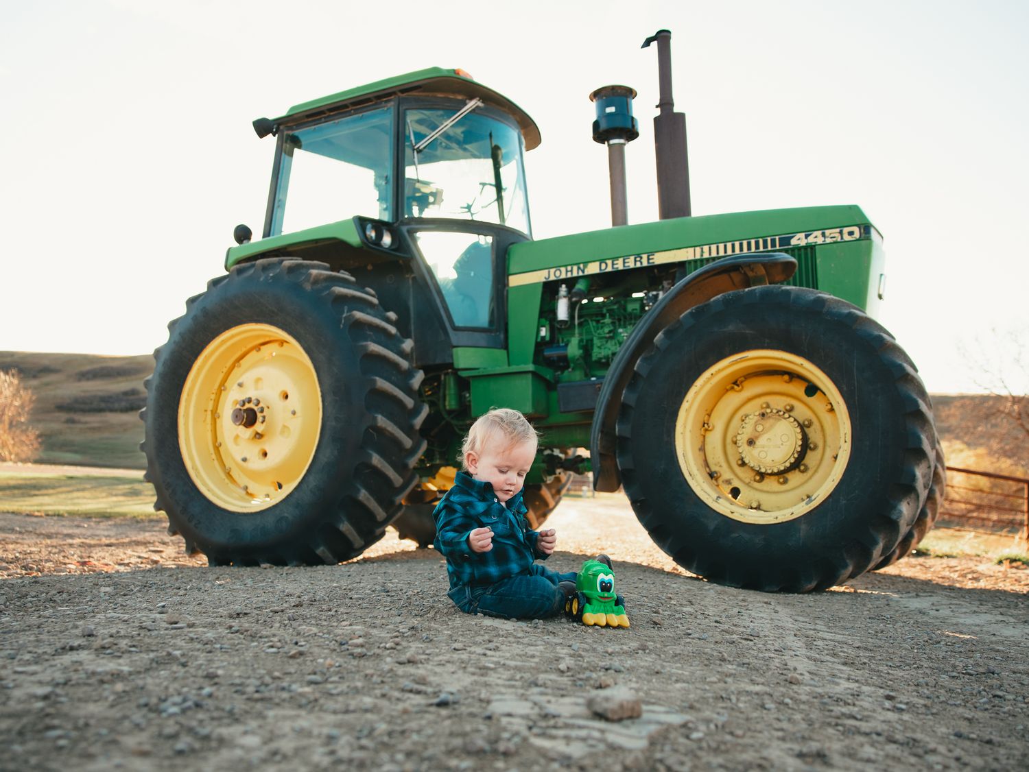 A baby sits on gravel playing with a toy tractor in front of a large green John Deere 4450 tractor.