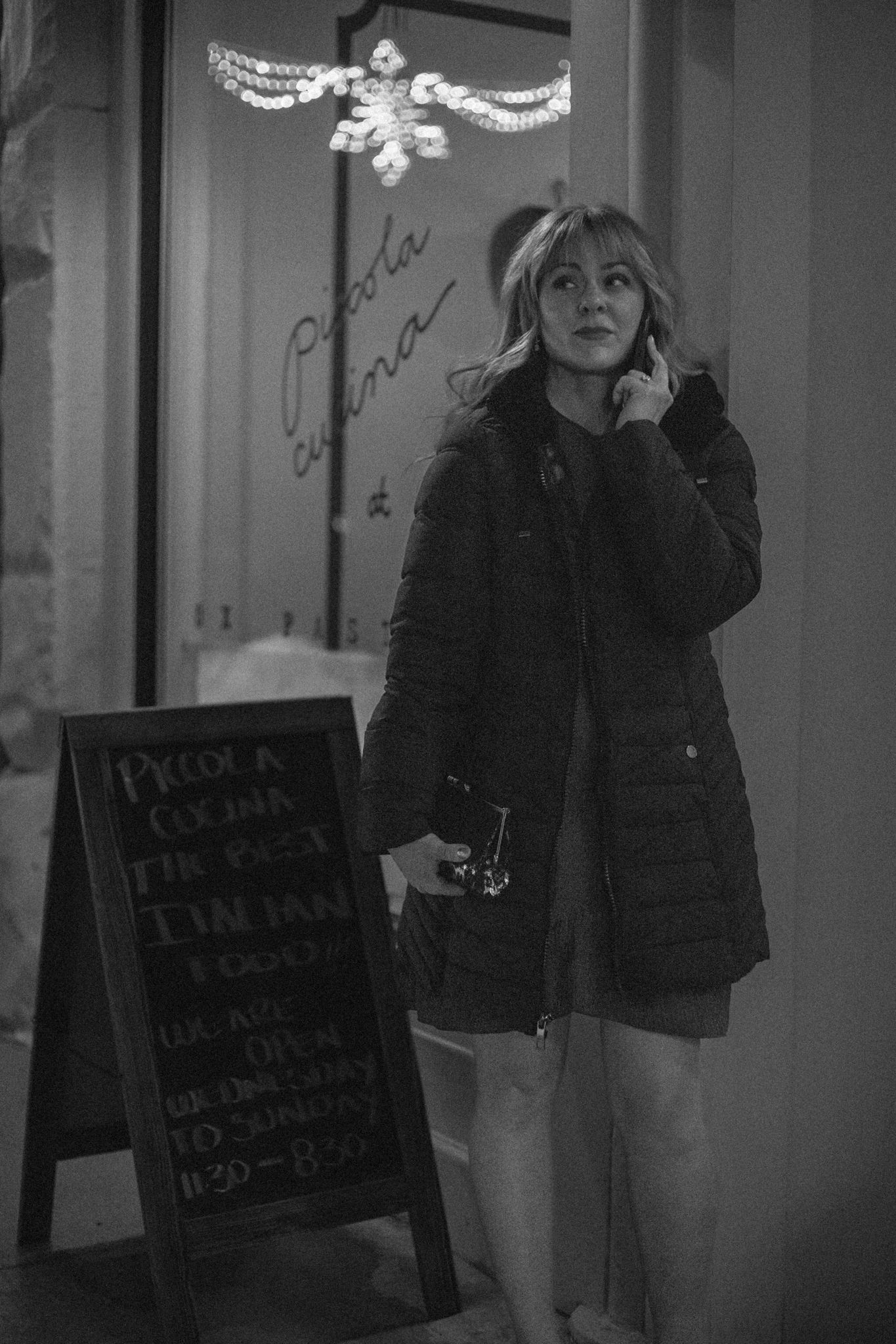 Person in a dark coat stands near chalkboard menu sign at night with holiday decorations visible overhead. Great Falls, Montana