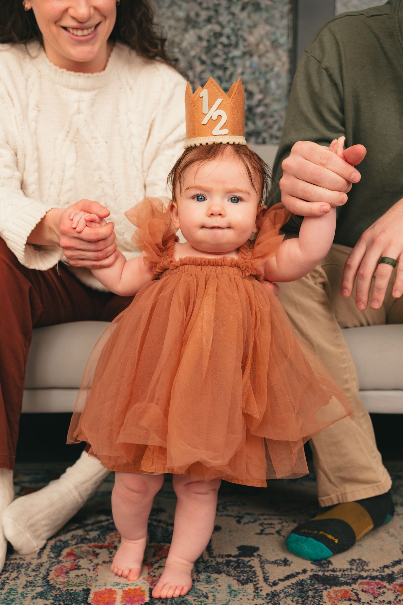 Baby girl in rust tulle dress wearing a half birthday crown stands with parents supporting her hands.