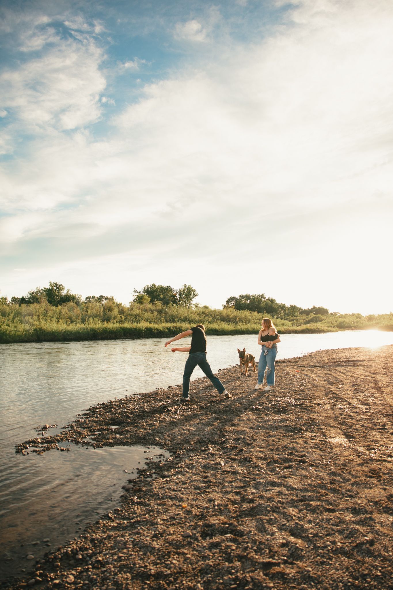 Two people and a dog walking along a rocky riverbank at golden hour with lush greenery in the background.