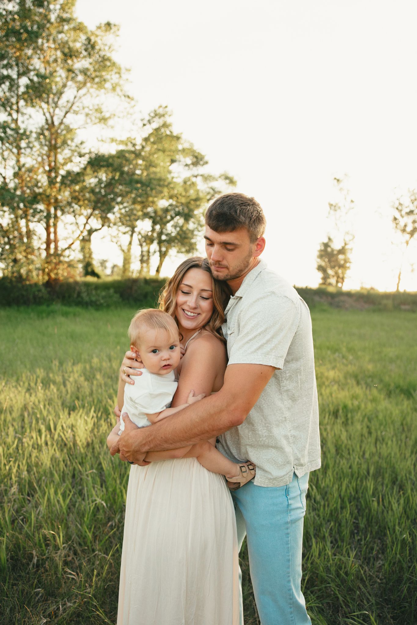 Young family with baby poses in golden hour sunlit green field, mother in white dress, father in light blue jeans.