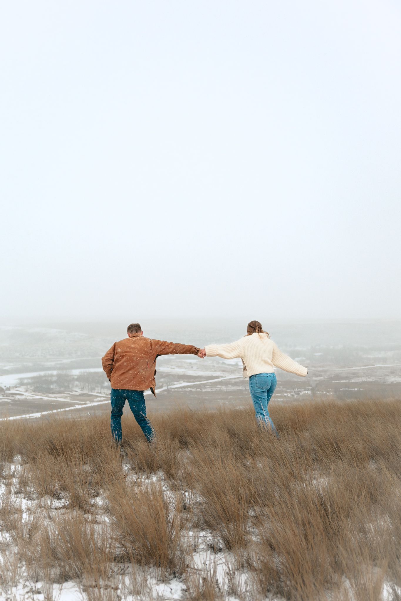 Couple holding hands running through snowy winter field on foggy day, viewed from behind.