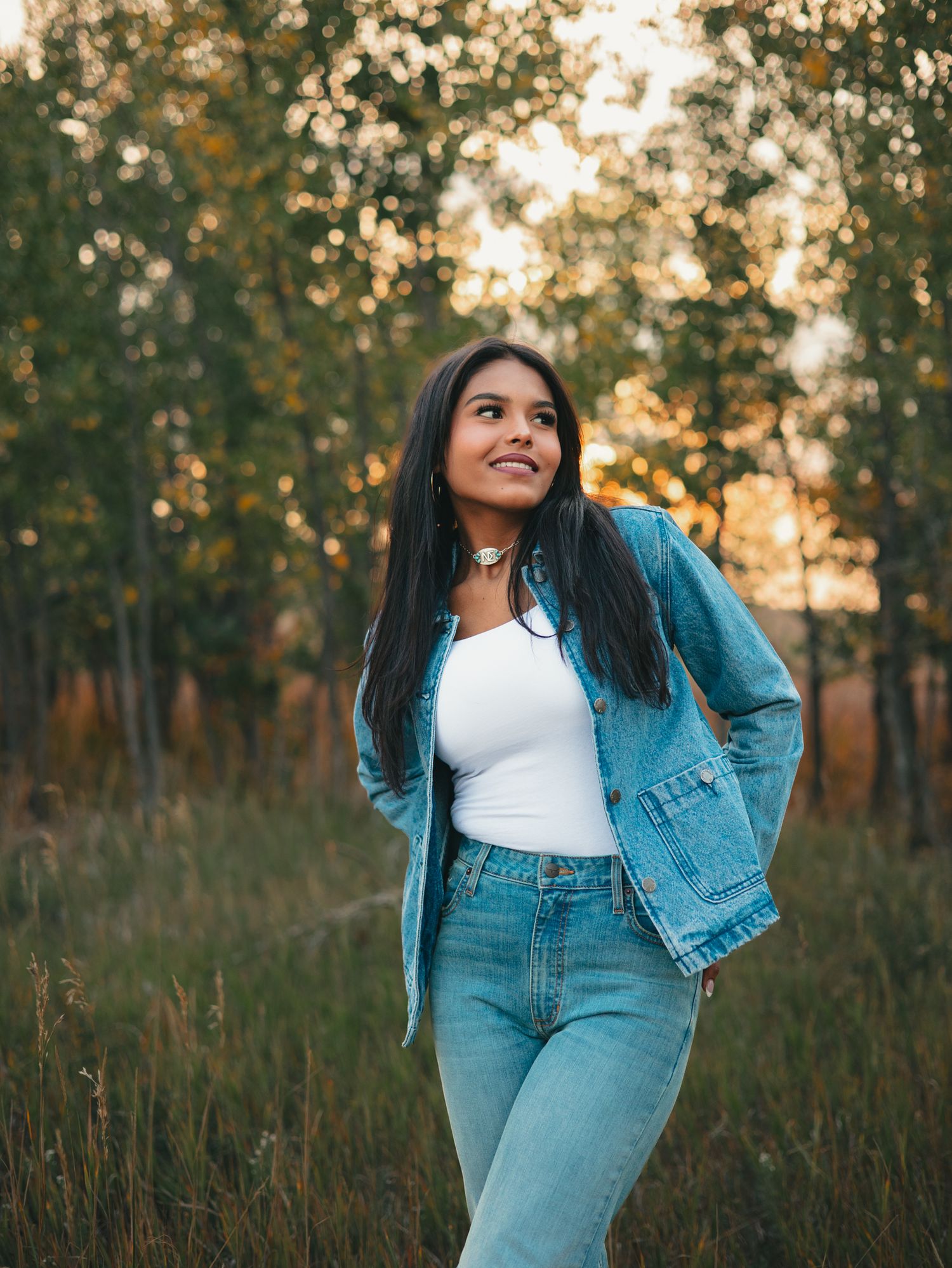 A senior in denim outfit poses in a field at golden hour with sunlit trees in the background. photo, Great Falls, MT
