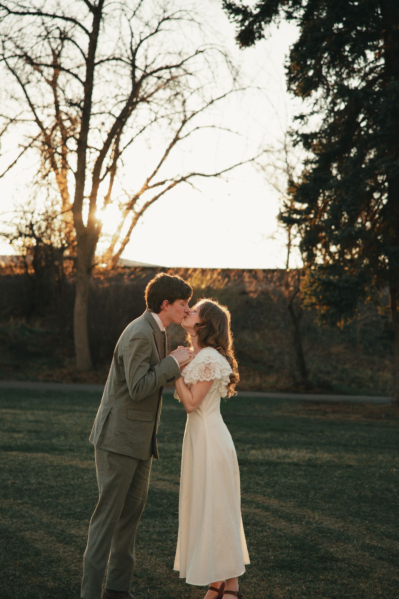 A couple shares a romantic kiss at golden hour sunset in a park, bride in white lace dress, groom in gray suit.