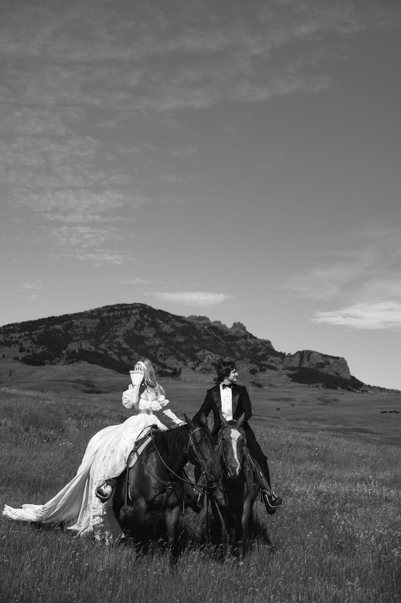 Black and white photo of a bride and groom riding horses in a field with mountains in the background.