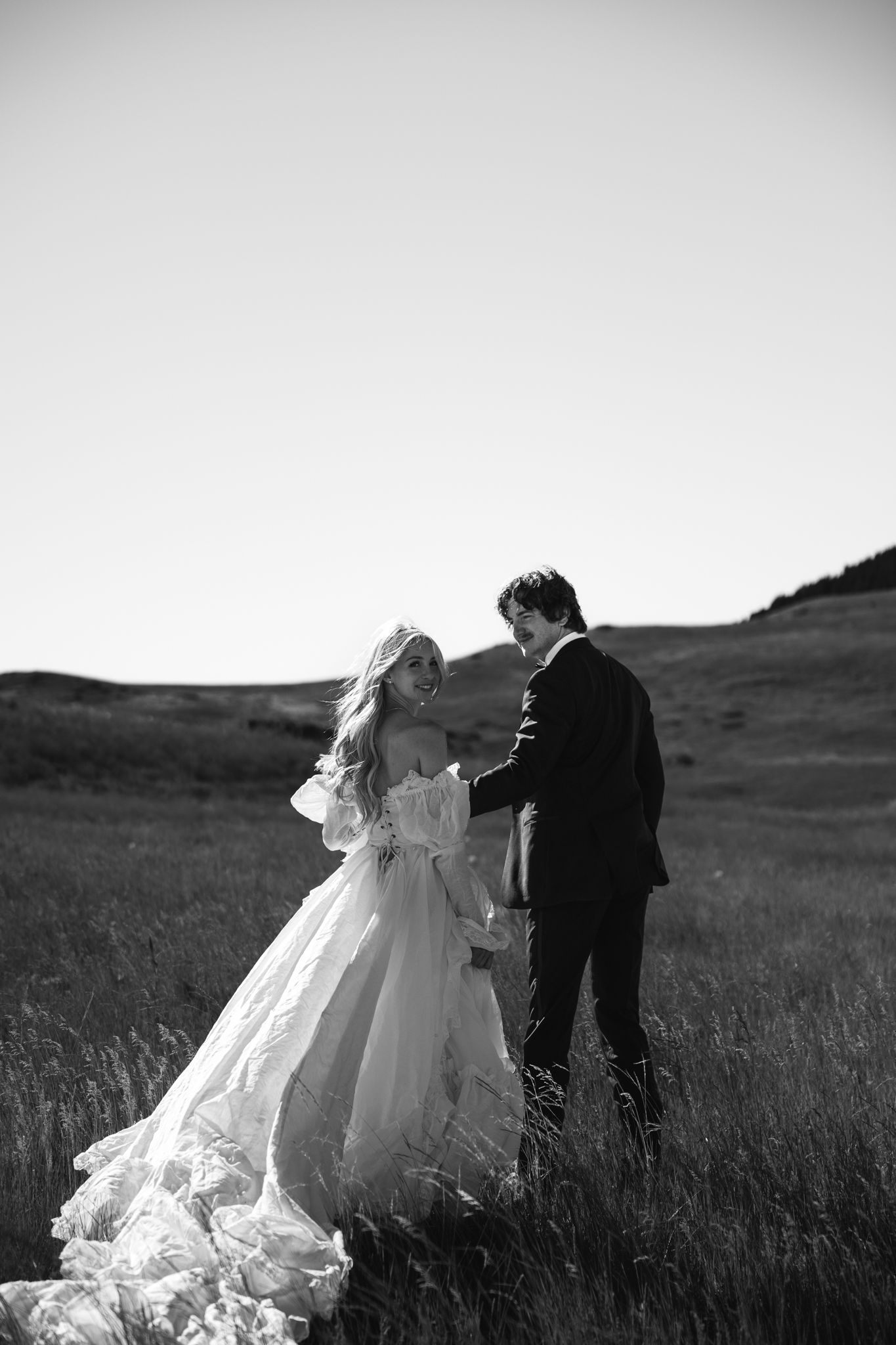 Black and white photo of bride in flowing gown and groom in suit holding hands in a scenic grassy field.
