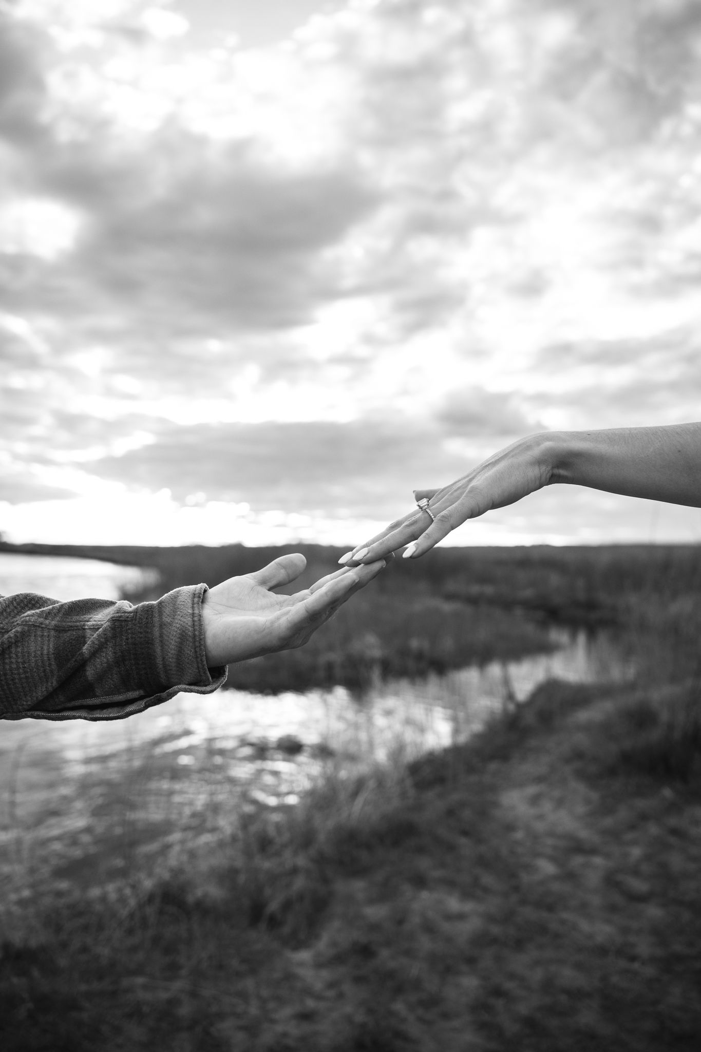 Black and white photo of two hands reaching toward each other against a cloudy sky and river landscape.