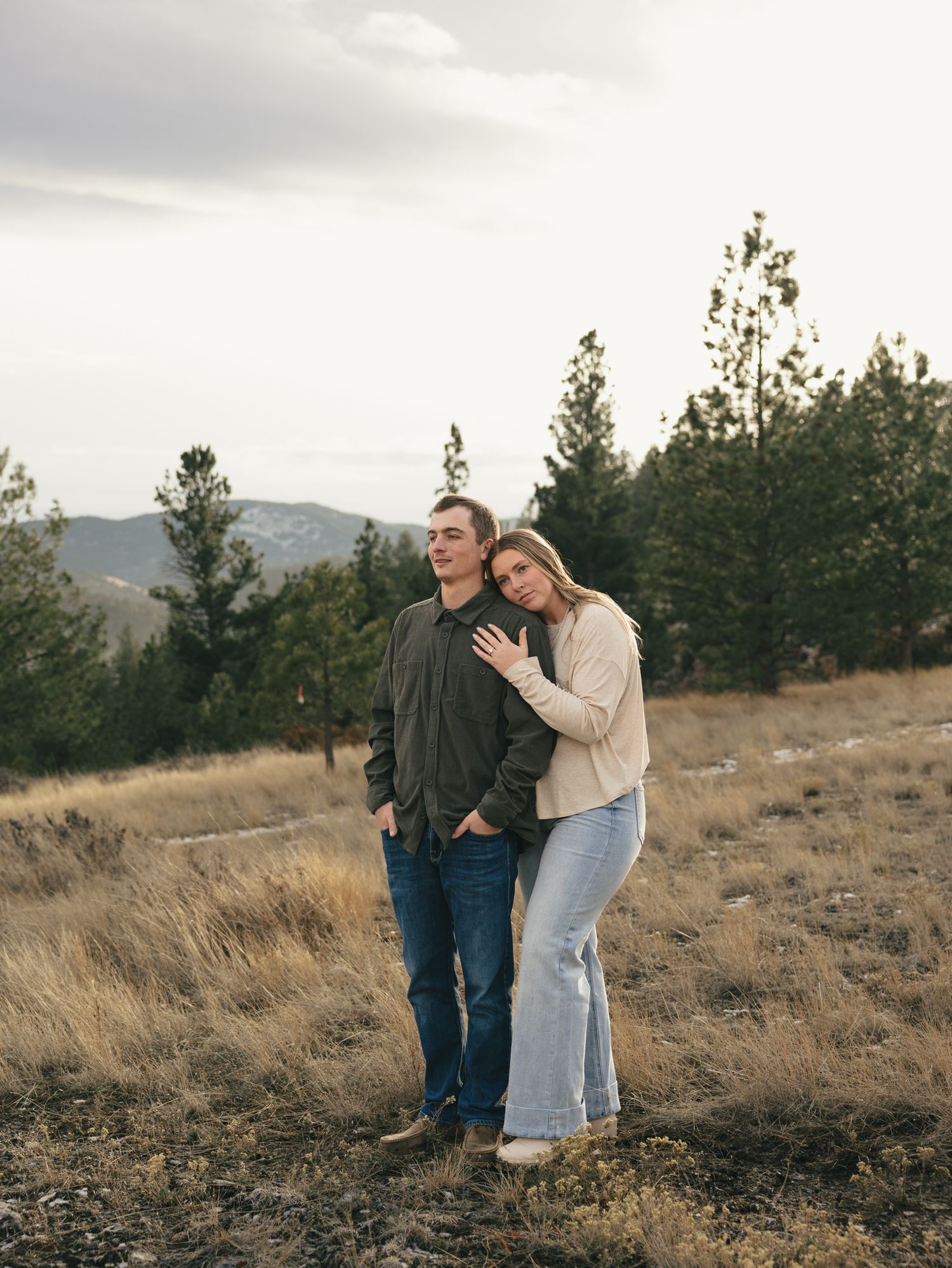 Couple embraces on a hillside with mountains and pine trees in the background during golden hour. engagement photo, Great Falls, Montana