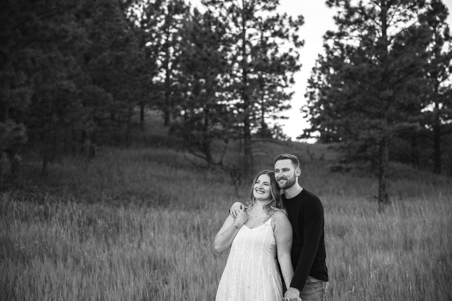 Black and white photo of a smiling couple standing in a field with tall grass and pine trees in the background.