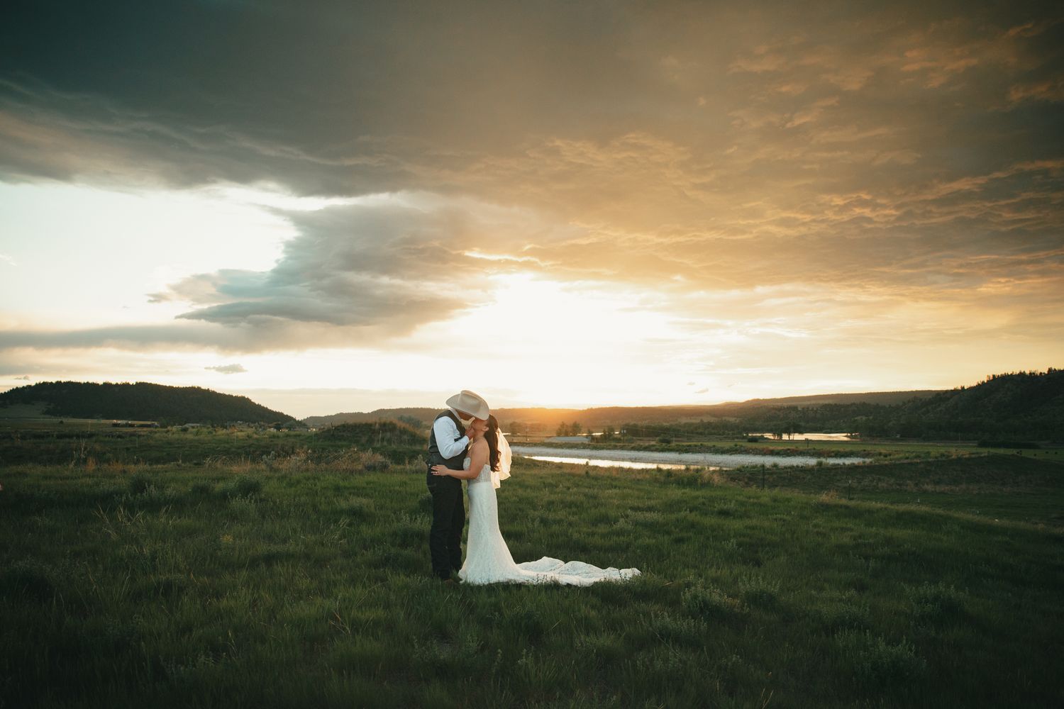Couple sharing a romantic kiss in a green field at golden sunset, bride in white gown with train, scenic countryside backdrop.