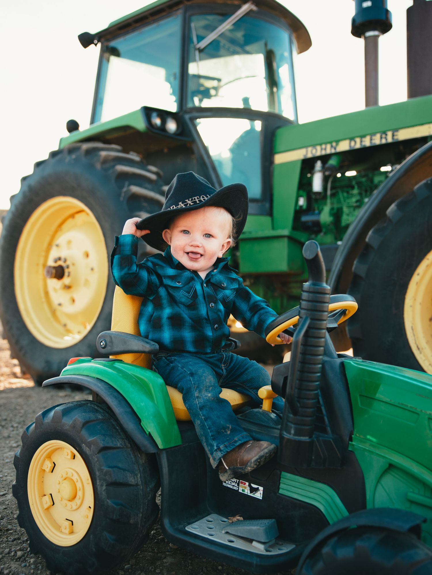 Smiling toddler in cowboy hat and plaid shirt sits on toy John Deere tractor with real tractor behind.