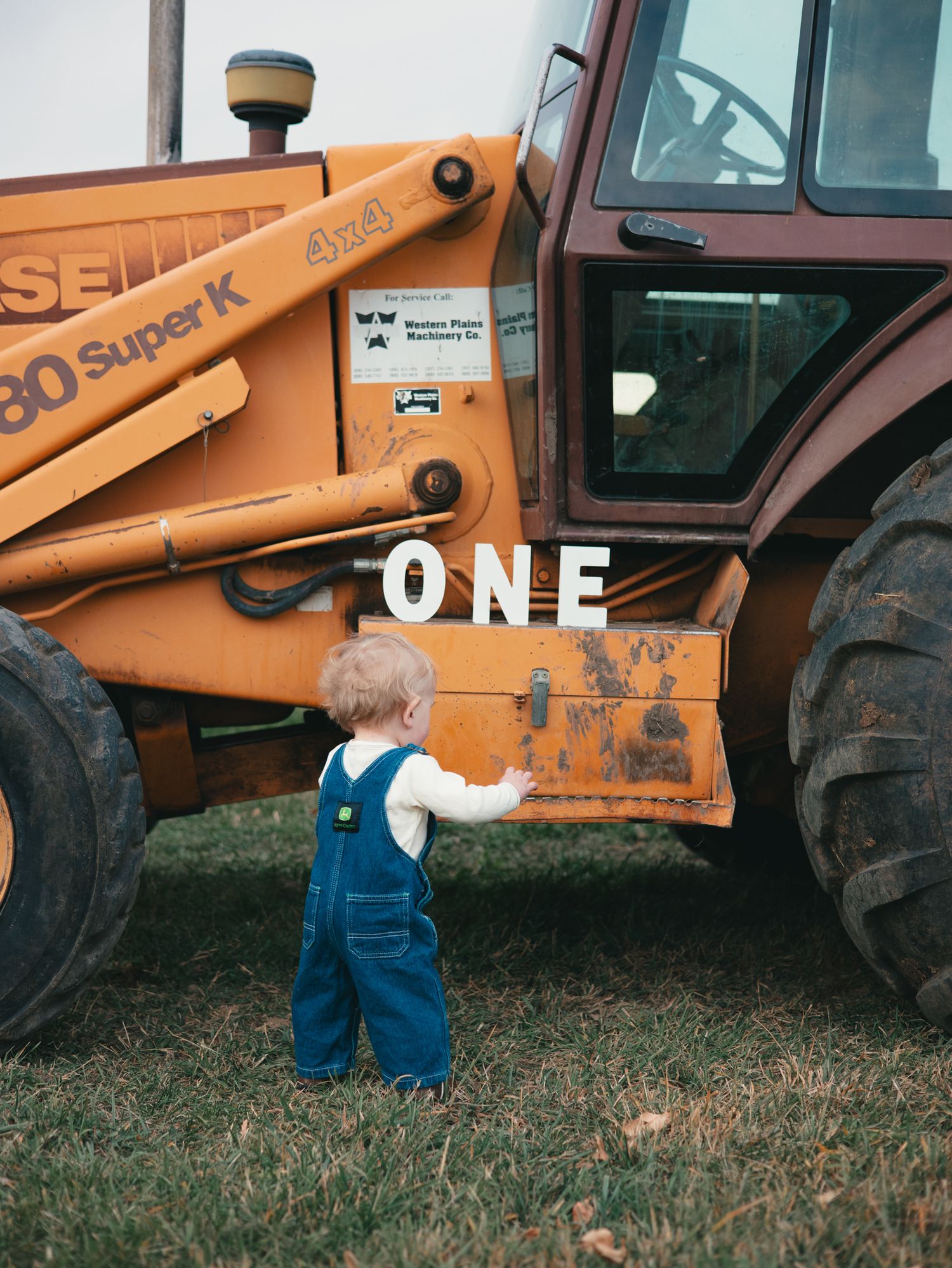 A toddler in denim overalls stands beside a large orange Case 480 Super K tractor with an ONE birthday sign.