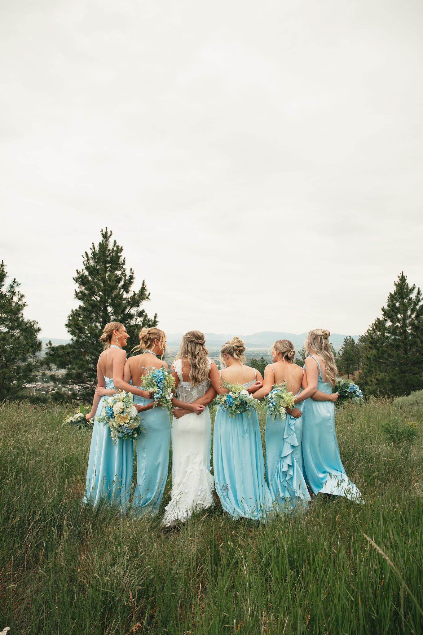 Bride and bridesmaids in light blue gowns holding floral bouquets, standing together in a lush green meadow with pine trees.